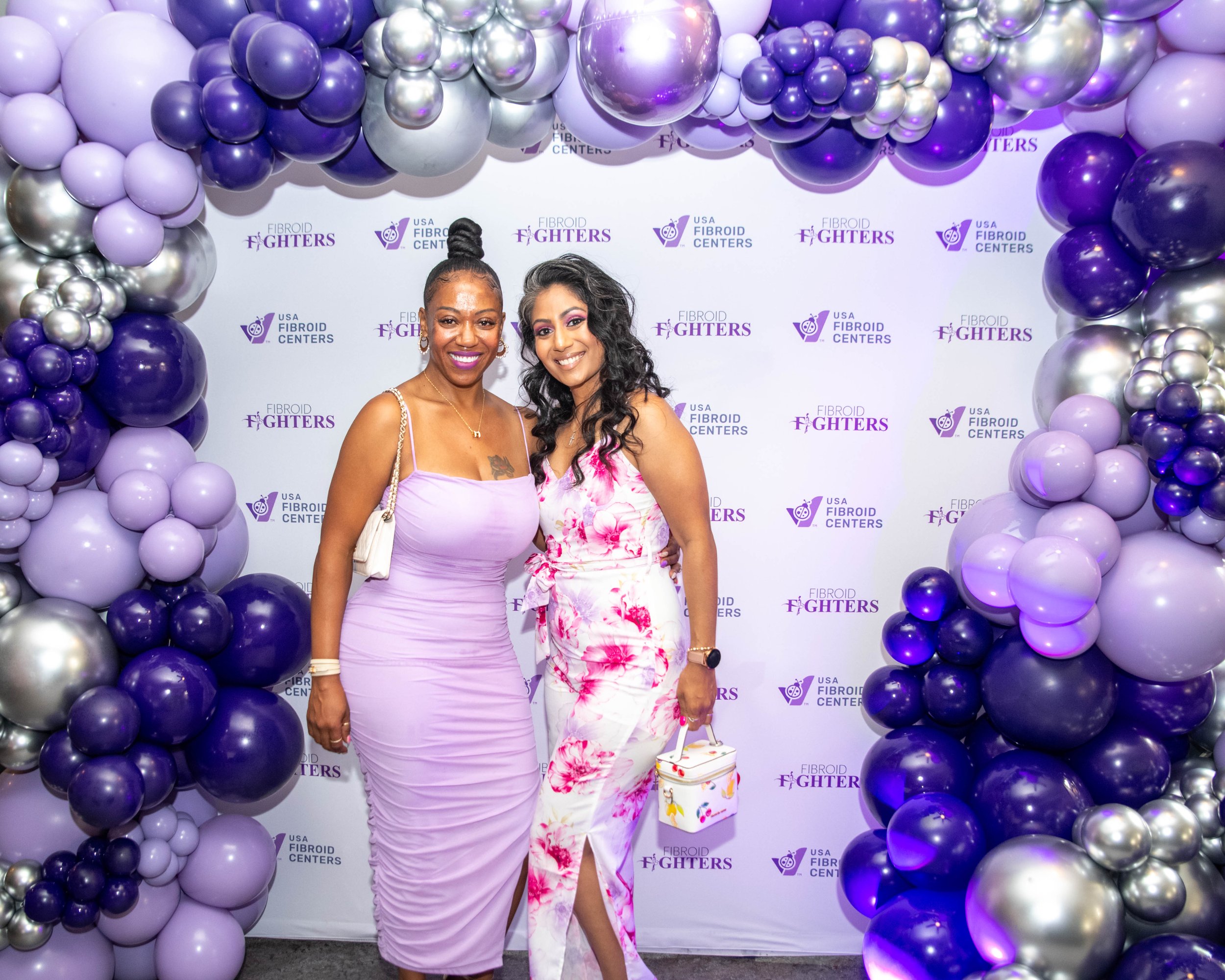 Two women standing together at a fundraising event, smiling, in front of a decorated backdrop with purple, lavender, and silver balloons. The backdrop has logos of the USA Fibroid Centers and Fibroid Fighters.