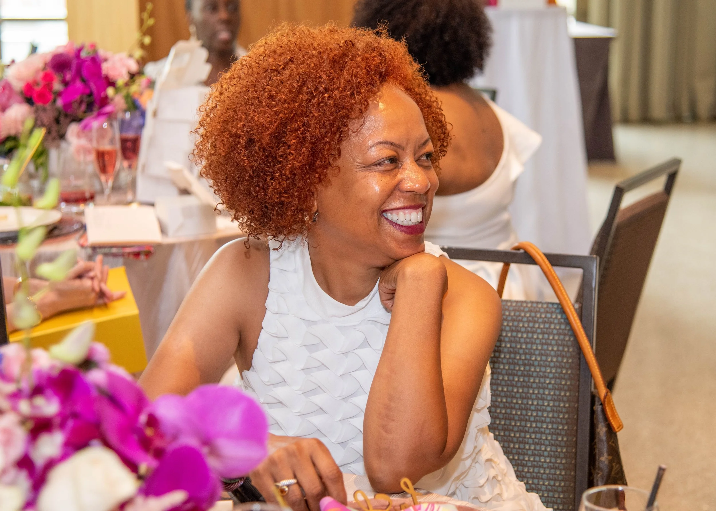 A woman with curly red hair and wearing a white ruffled dress smiling at a social gathering or celebration.
