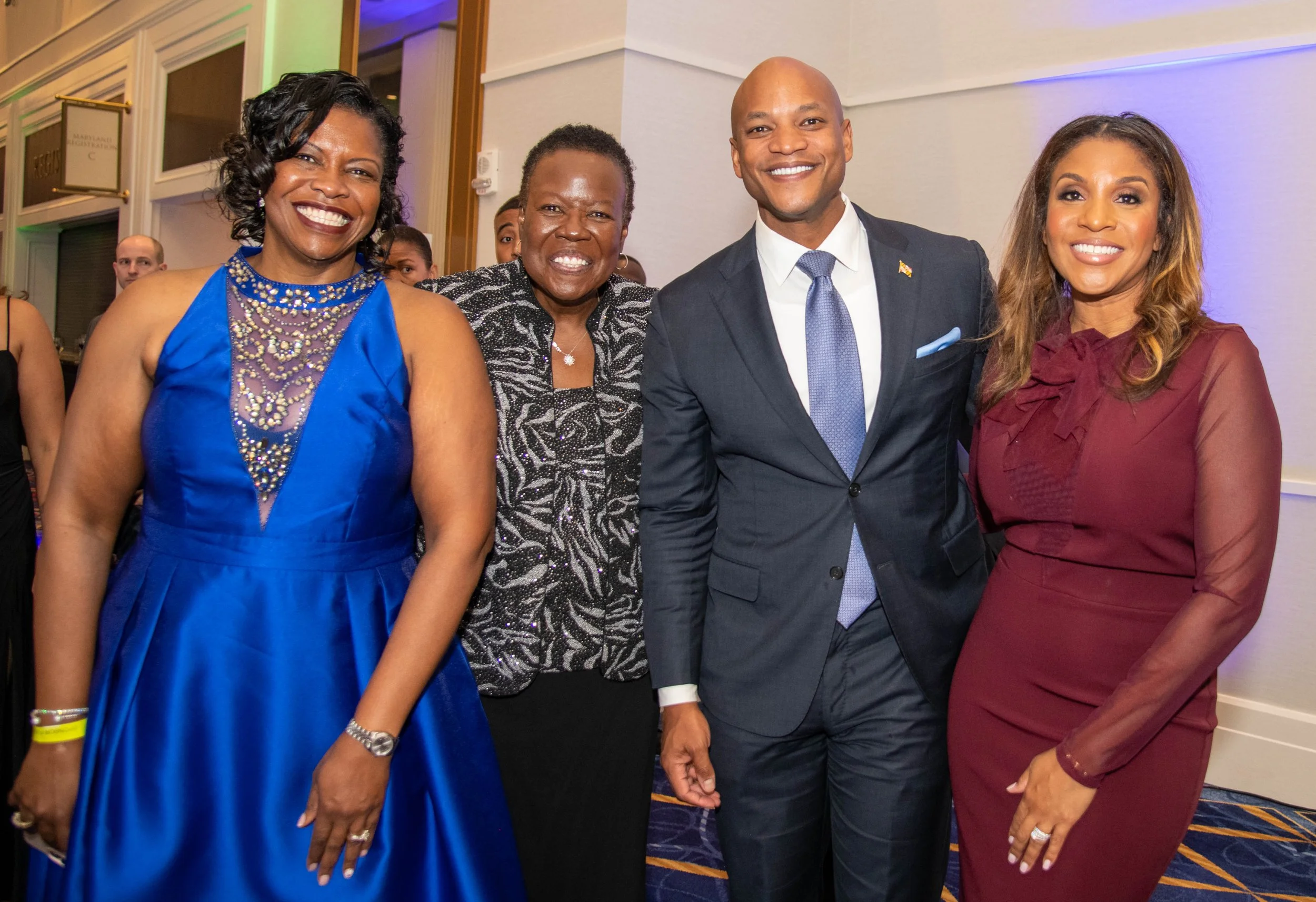 Group of four people wearing formal attire, smiling for the camera, at an indoor social event with colorful lighting.