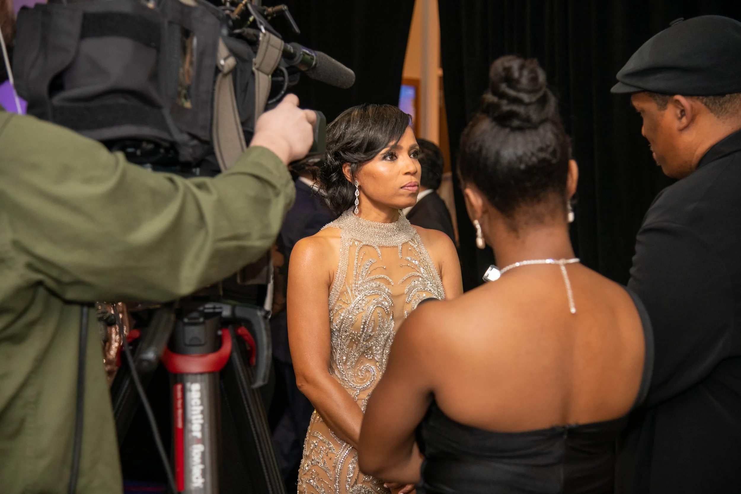 A woman in an elegant, beige evening gown being interviewed or filmed by a crew, with a camera operator in the foreground and two people in conversation, all dressed formally.