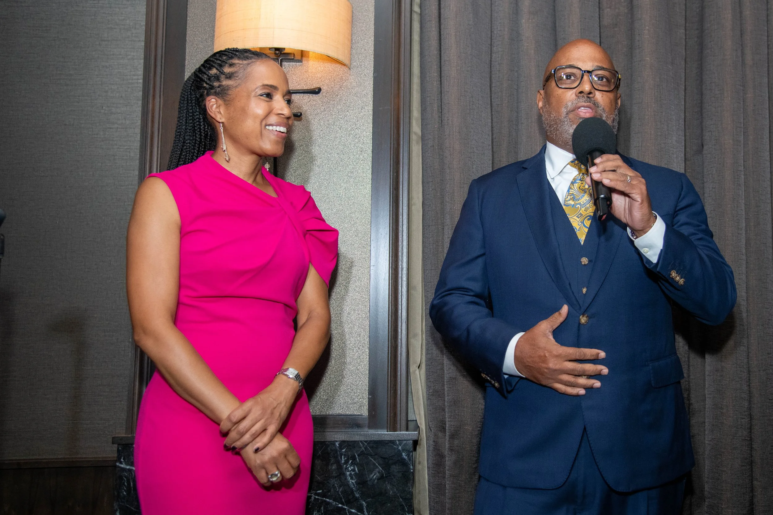 A woman in a bright pink dress smiling and looking at a man in a navy suit who is speaking into a microphone.
