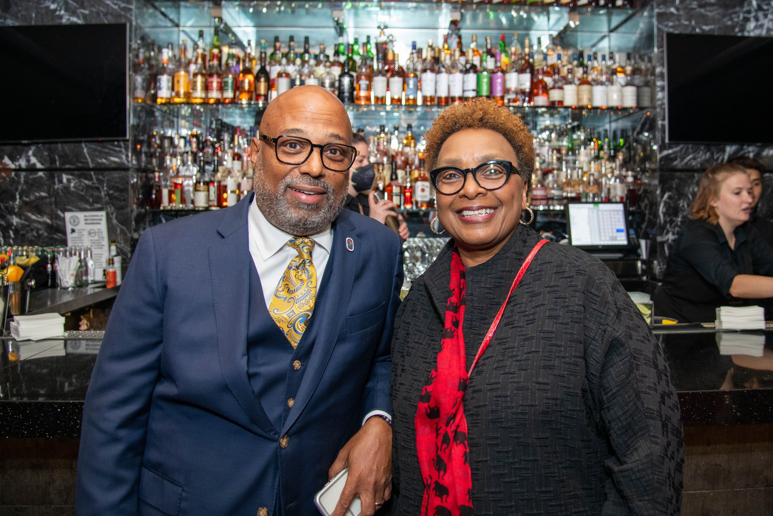 Two people, a man and a woman, smiling at the camera in a bar or restaurant setting, with a variety of liquor bottles displayed on shelves behind them.