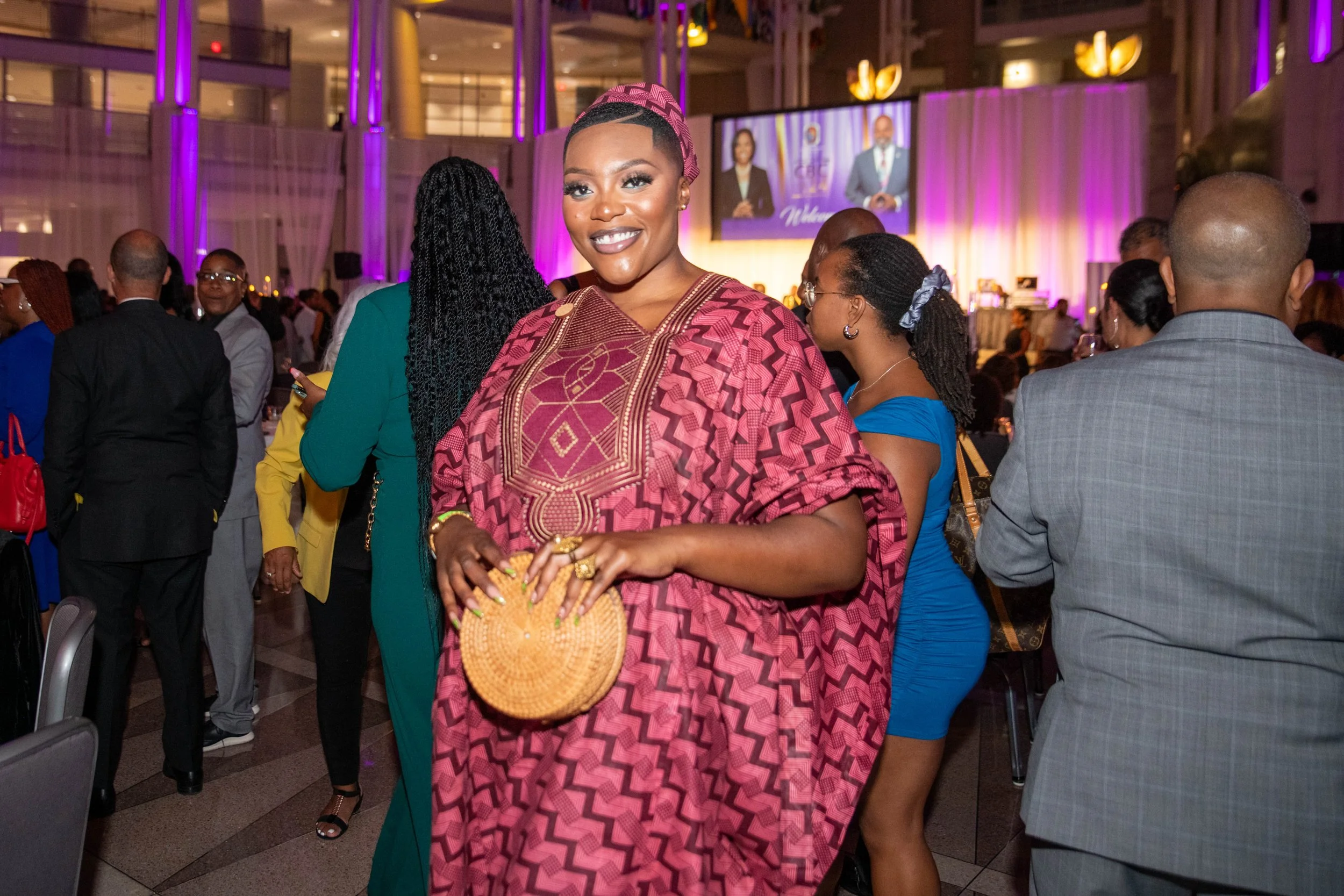 A woman in traditional African attire smiling at an event, holding a woven clutch bag, with a lively crowd and stage in the background.