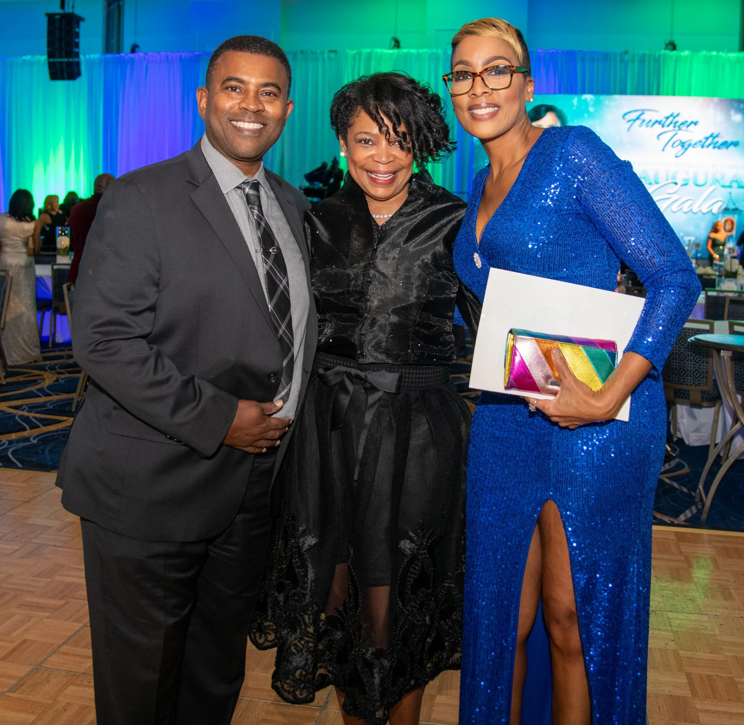 Three well-dressed people smiling at an indoor event with a colorful background. The person on the right is wearing a bright blue sequin dress, holding a rainbow-colored purse and a white envelope. The woman in the middle is wearing a black lace dres
