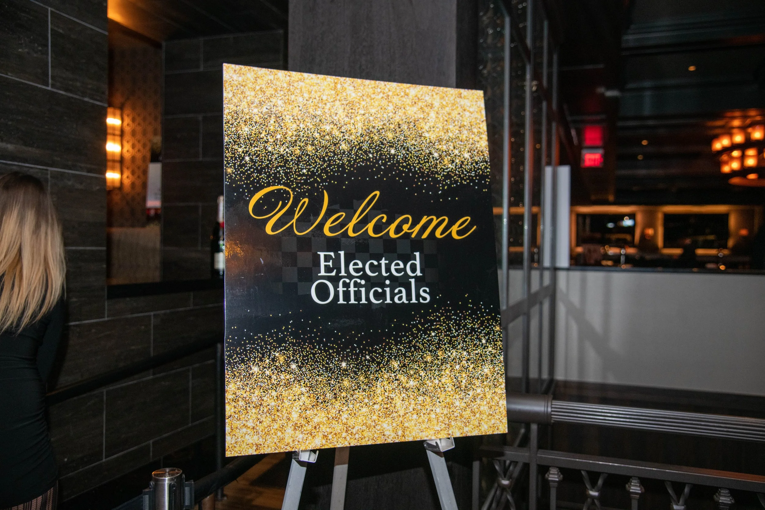 Sign welcoming elected officials decorated with gold glitter design in a restaurant or event space.