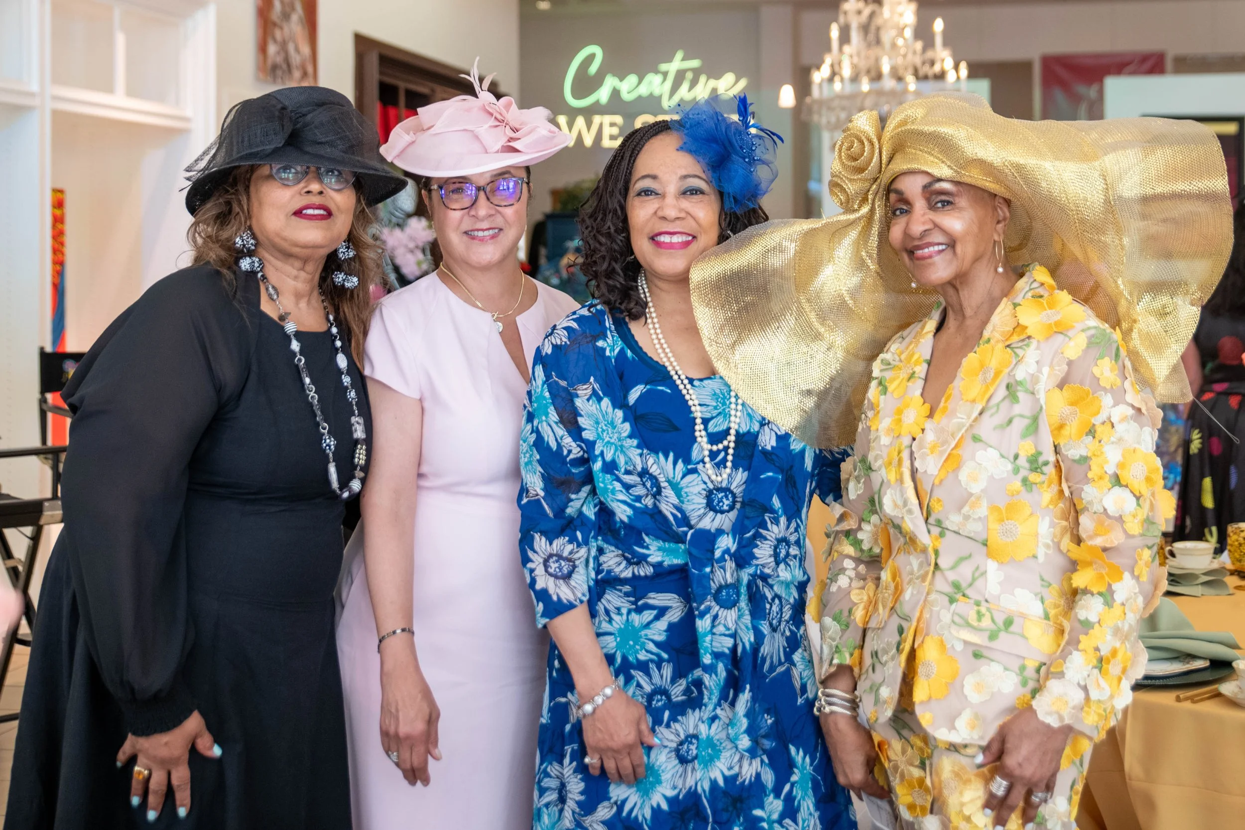 Four women dressed in colorful, fashionable outfits, wearing elaborate hats, standing together and smiling at an indoor event.