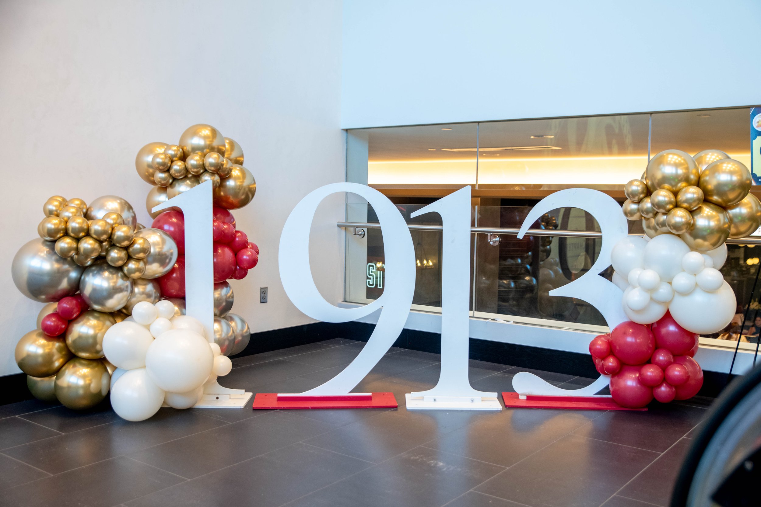 Large white 2013 numbers with balloon arrangements on each side, featuring gold, white, red, and silver balloons, displayed indoors against a wall.