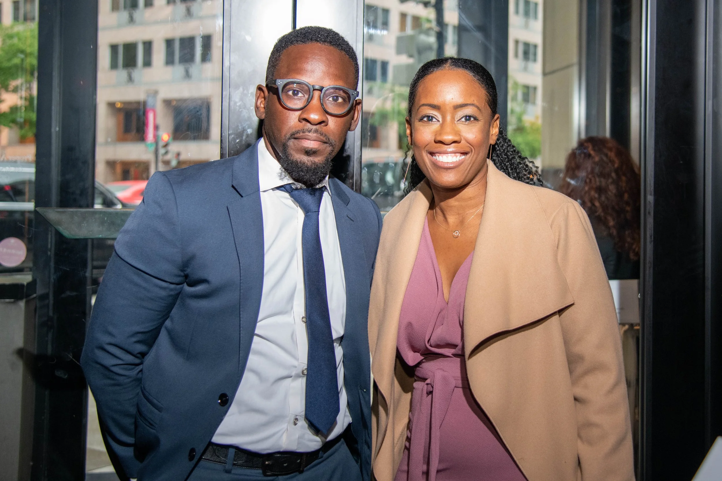 A man and a woman standing indoors near large glass doors, dressed in business attire, smiling at the camera.