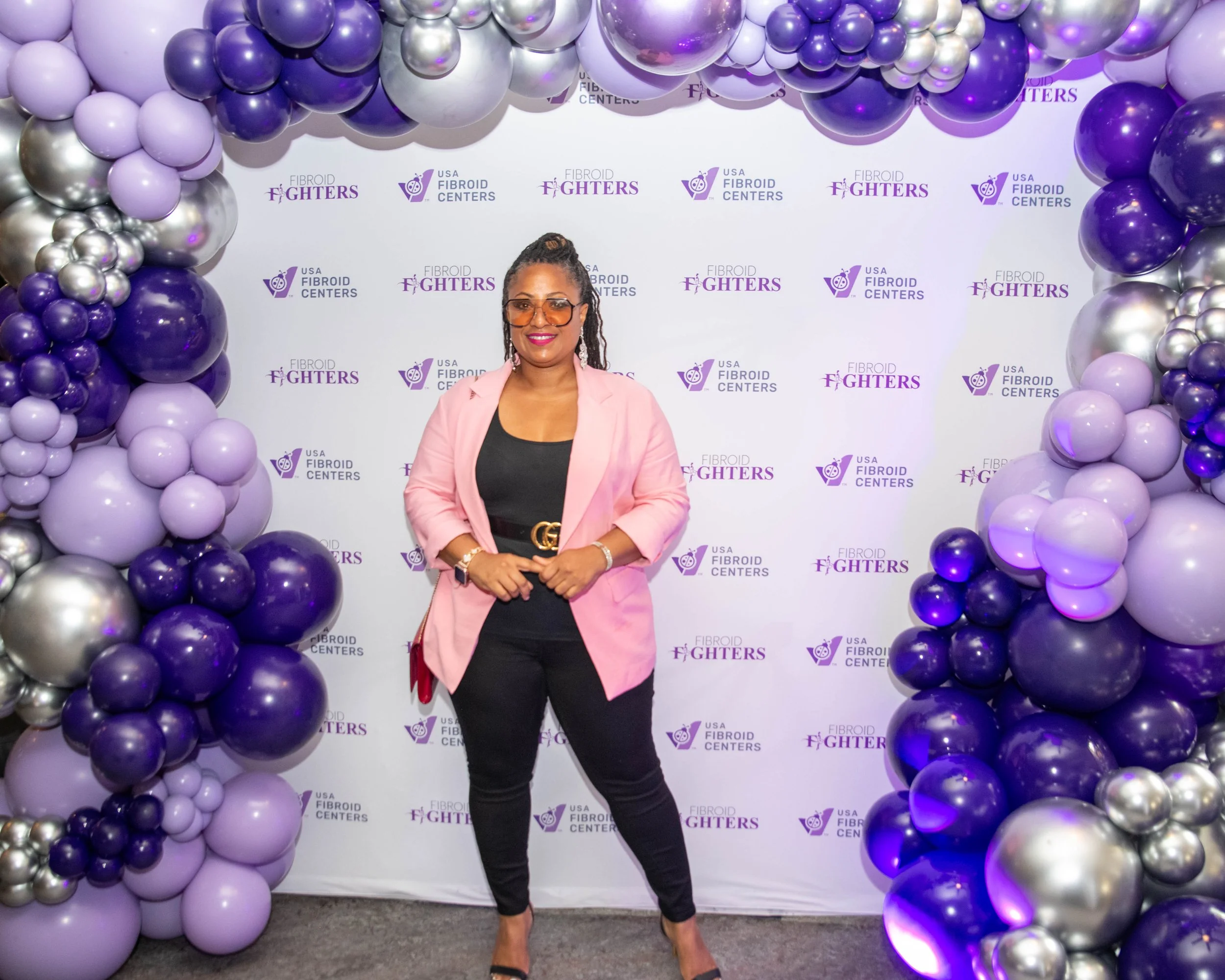 A woman standing in front of a backdrop with a purple and silver balloon arch. She is wearing a pink blazer, black top and pants, and sunglasses, smiling for the camera at a USA Fibroid Centers event.