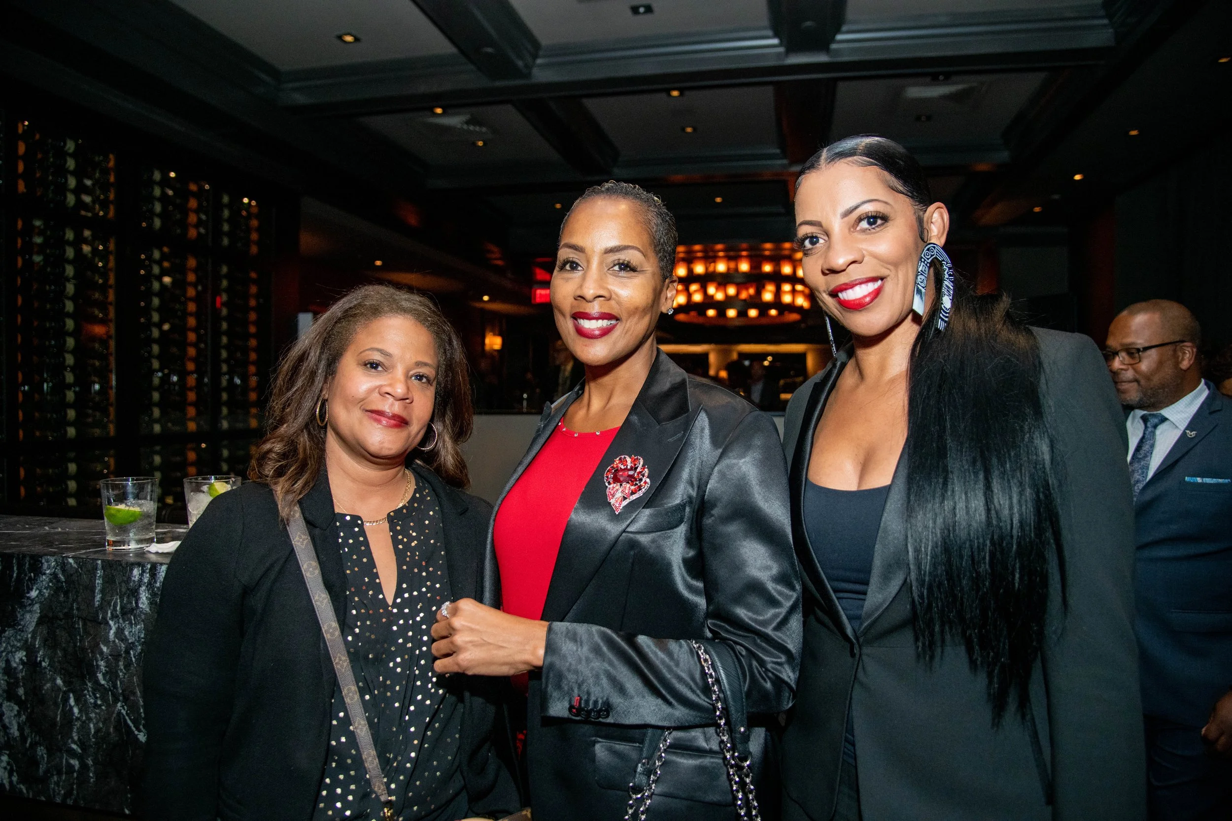 Three women smiling and posing together at an indoor event. They are dressed in formal attire, with the woman on the right wearing blue earrings and the woman in the middle wearing a brooch. The background features warm lighting and a bar area.