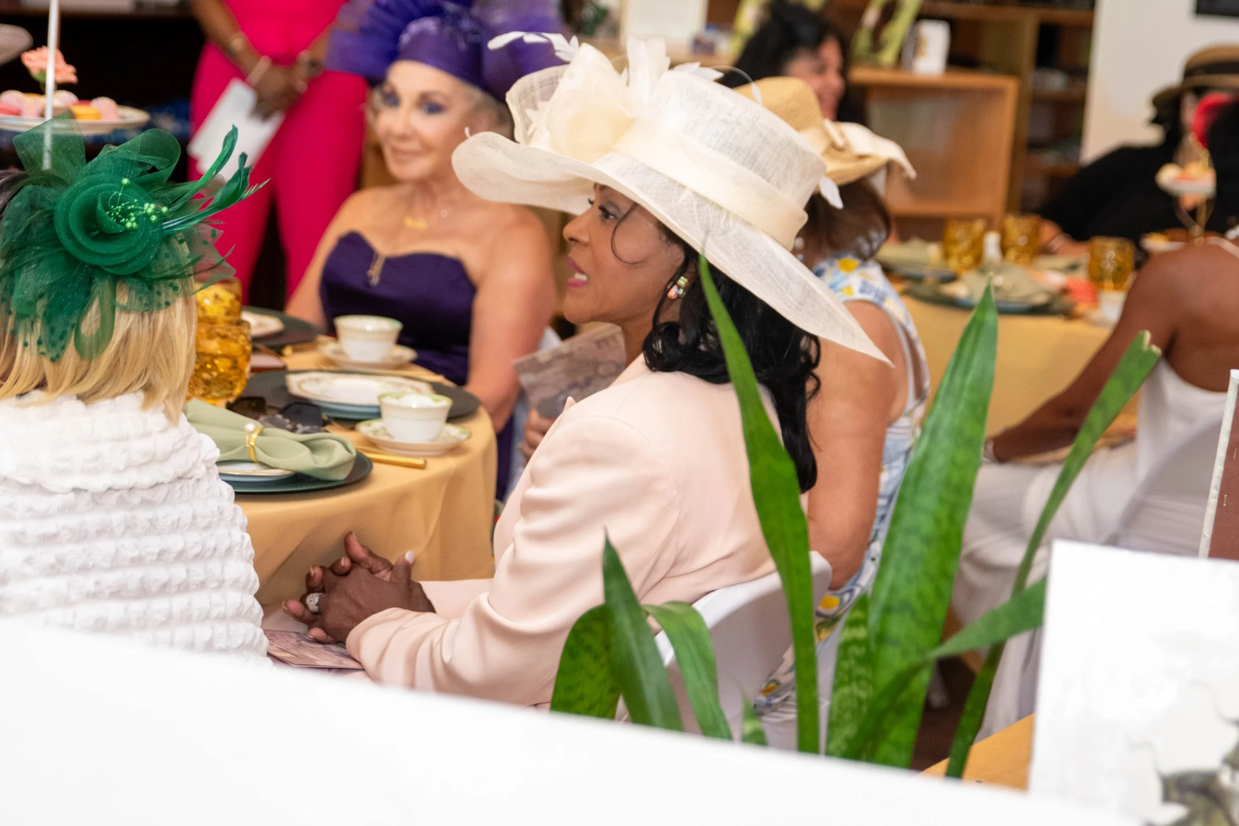 Women wearing elegant hats and dresses sitting around a dining table during a social gathering or celebration.