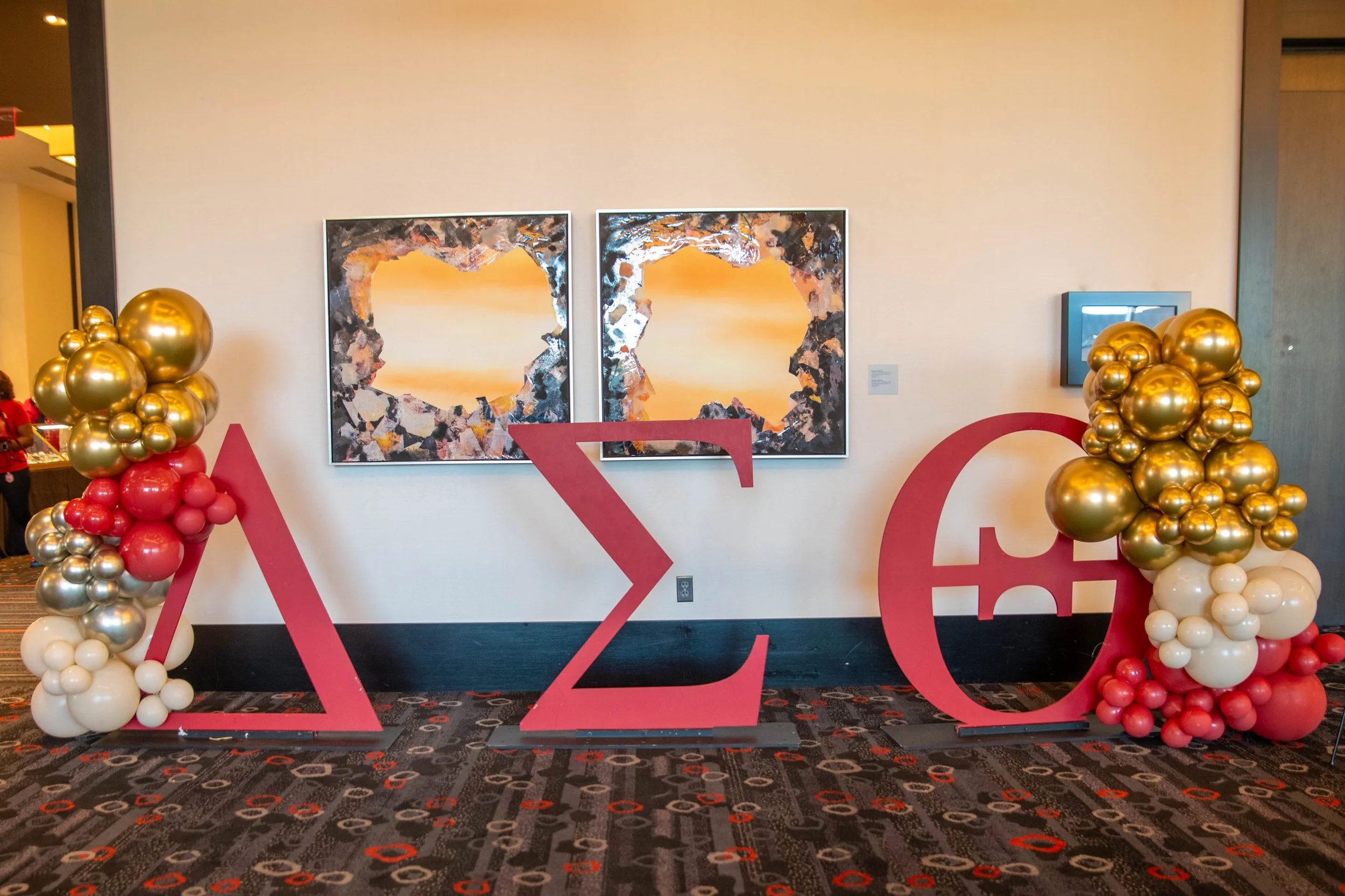 Decorative display with large red Greek letters Lambda, Sigma, and Epsilon, surrounded by balloon arrangements in gold, red, and white, with two abstract paintings featuring sunset scenes in the background.