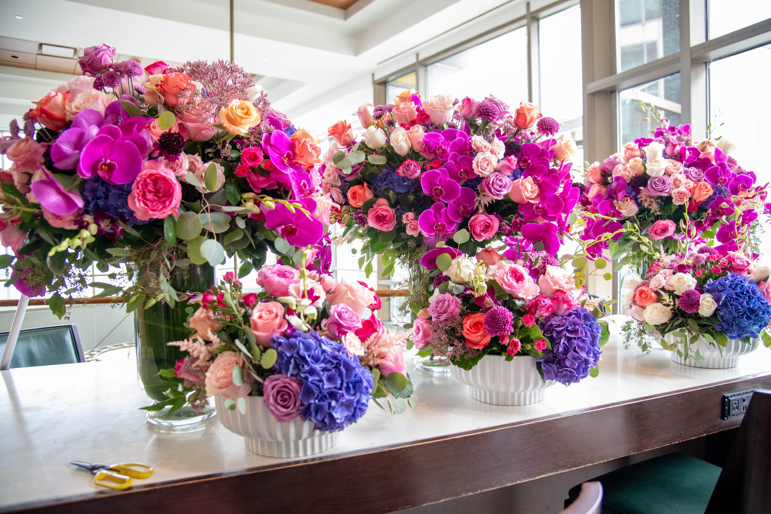 Multiple large floral arrangements with pink, purple, peach, and white flowers on a table near windows.