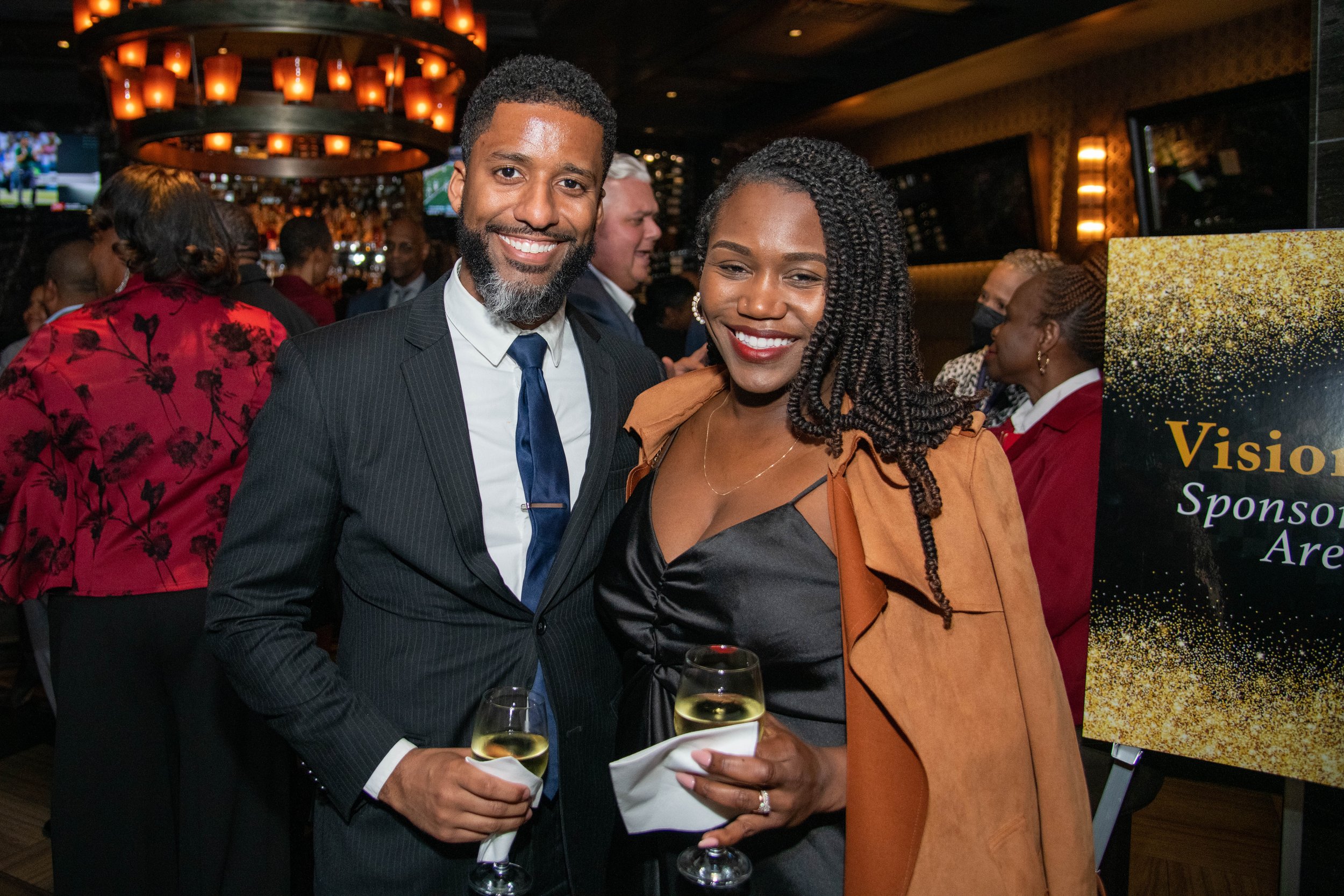 Two people, a man in a suit with a tie and a woman in a black dress with an orange jacket, smiling and holding glasses of white wine at a social event with many other attendees in the background.