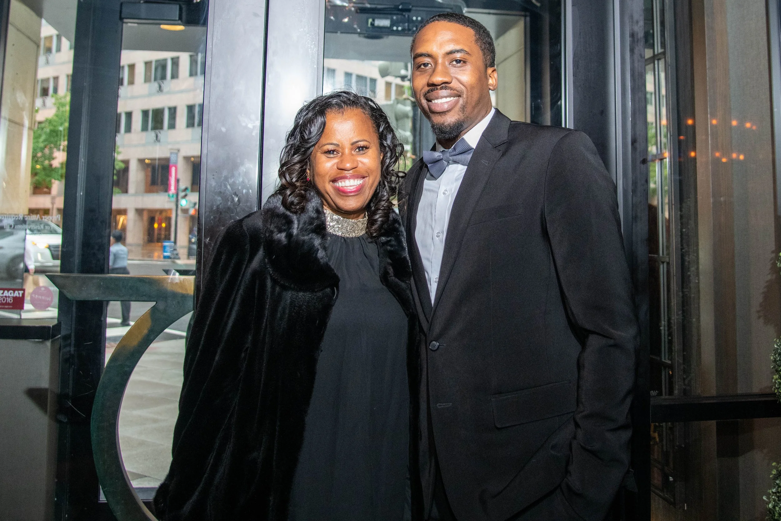 A smiling woman and man in formal clothing, standing together inside near a glass door with an city street outside in the background.
