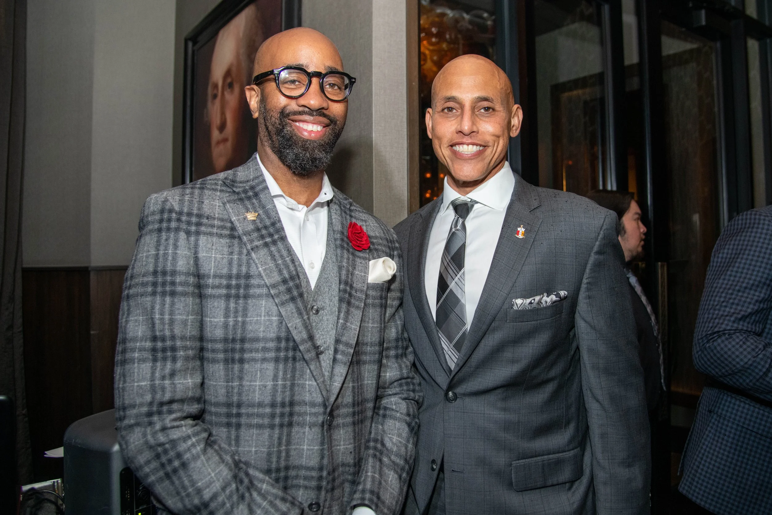 Two men in suits standing together at an indoor event, smiling at the camera. One man has a plaid gray suit, glasses, and a red lapel pin, the other in a dark gray suit with a patterned tie and pocket square.
