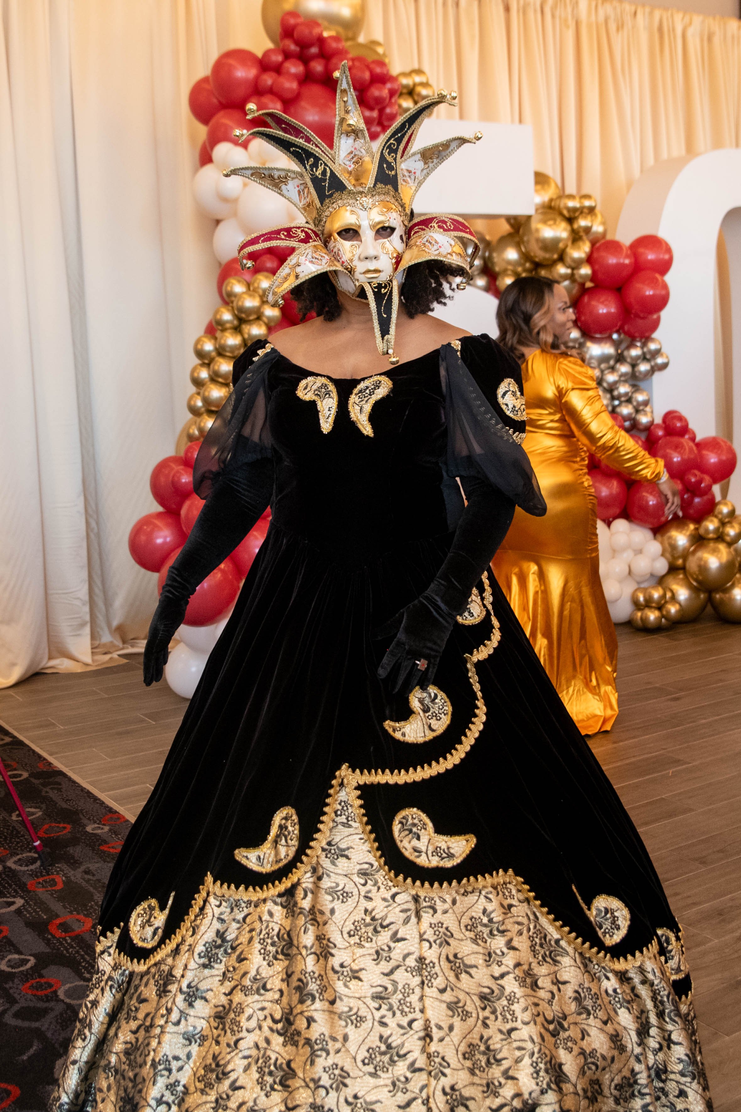 Person dressed in a black and gold velvet gown with ornate embroidery, wearing an elaborate Venetian-style mask with gold and black details, and a large jester-like headdress with sharp points and gold accents, standing in front of a festive balloon 