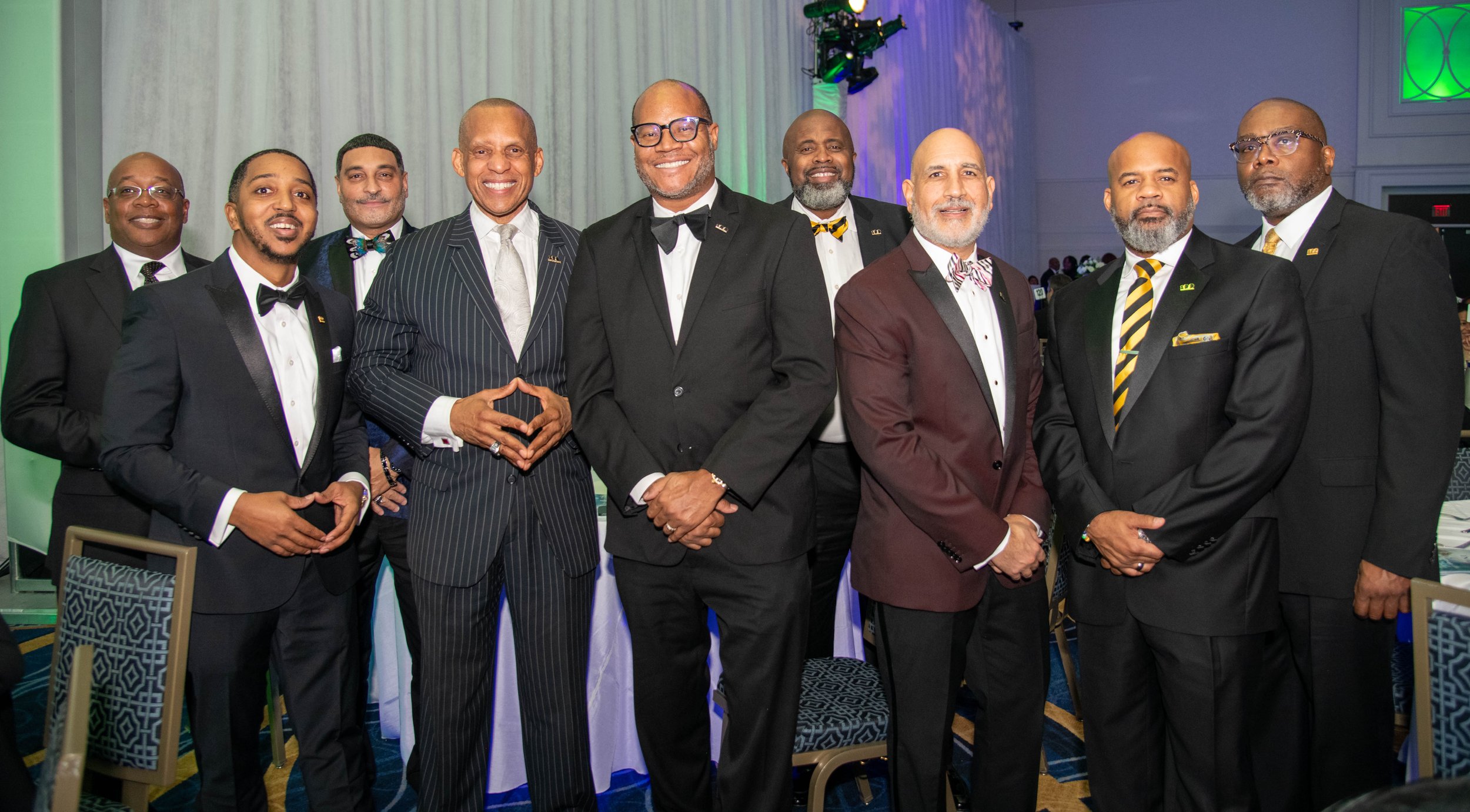 Group of men in formal suits and bow ties at an indoor event, smiling for the camera.