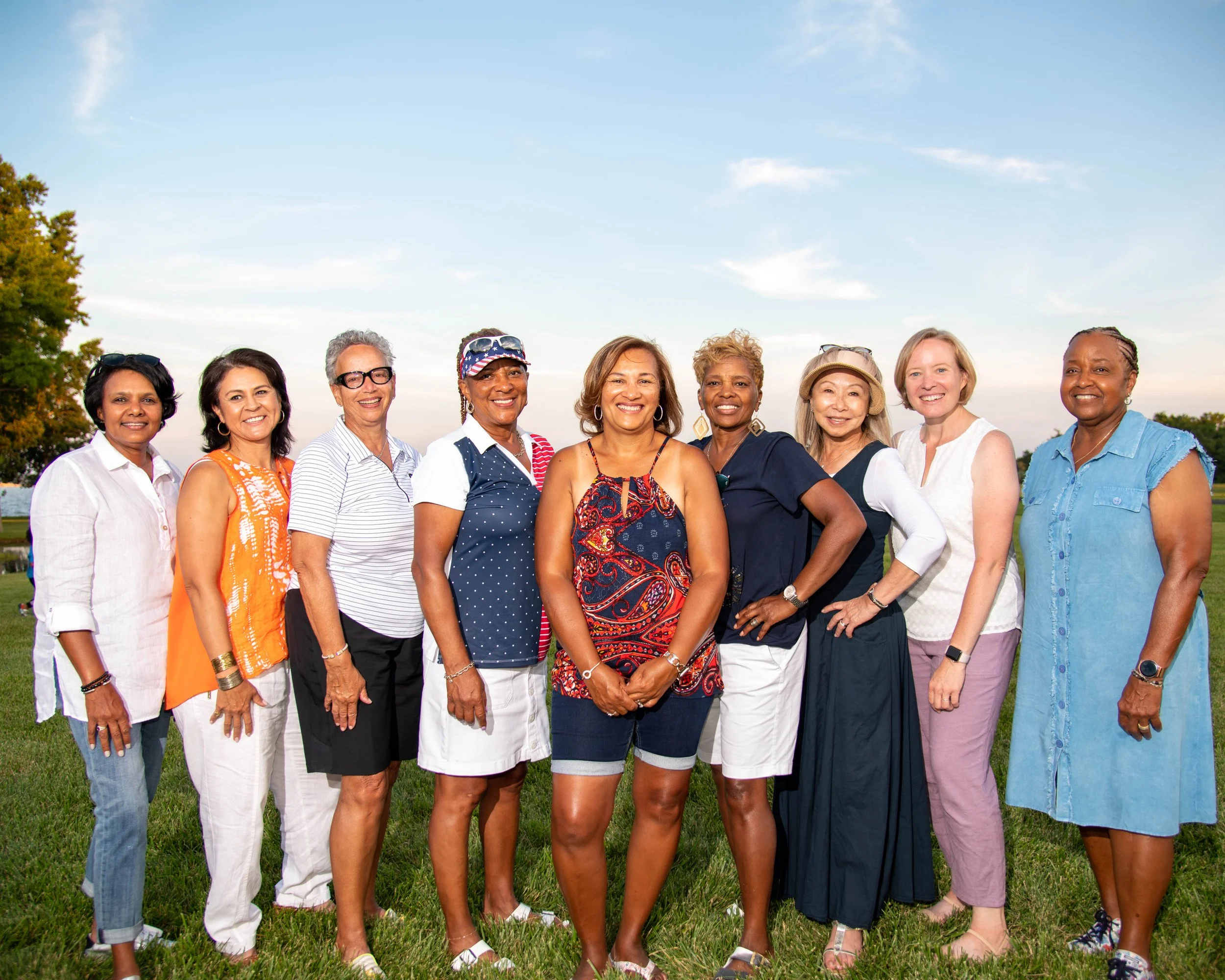 A group of ten women standing outdoors on grass, smiling at the camera, with trees and a blue sky in the background.