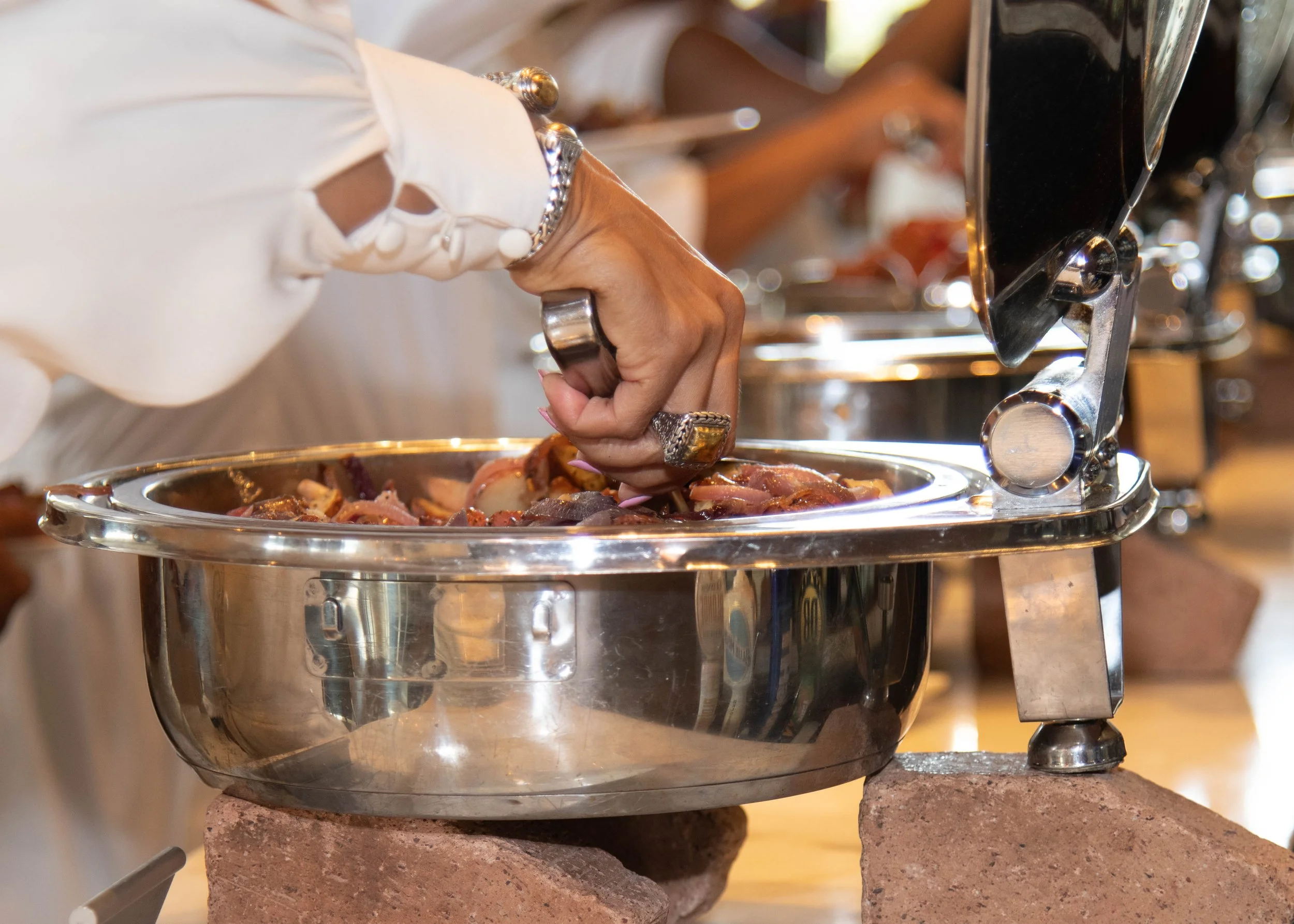 Person serving food from a stainless steel chafing dish at a buffet, with a close-up view of their hand wearing rings.