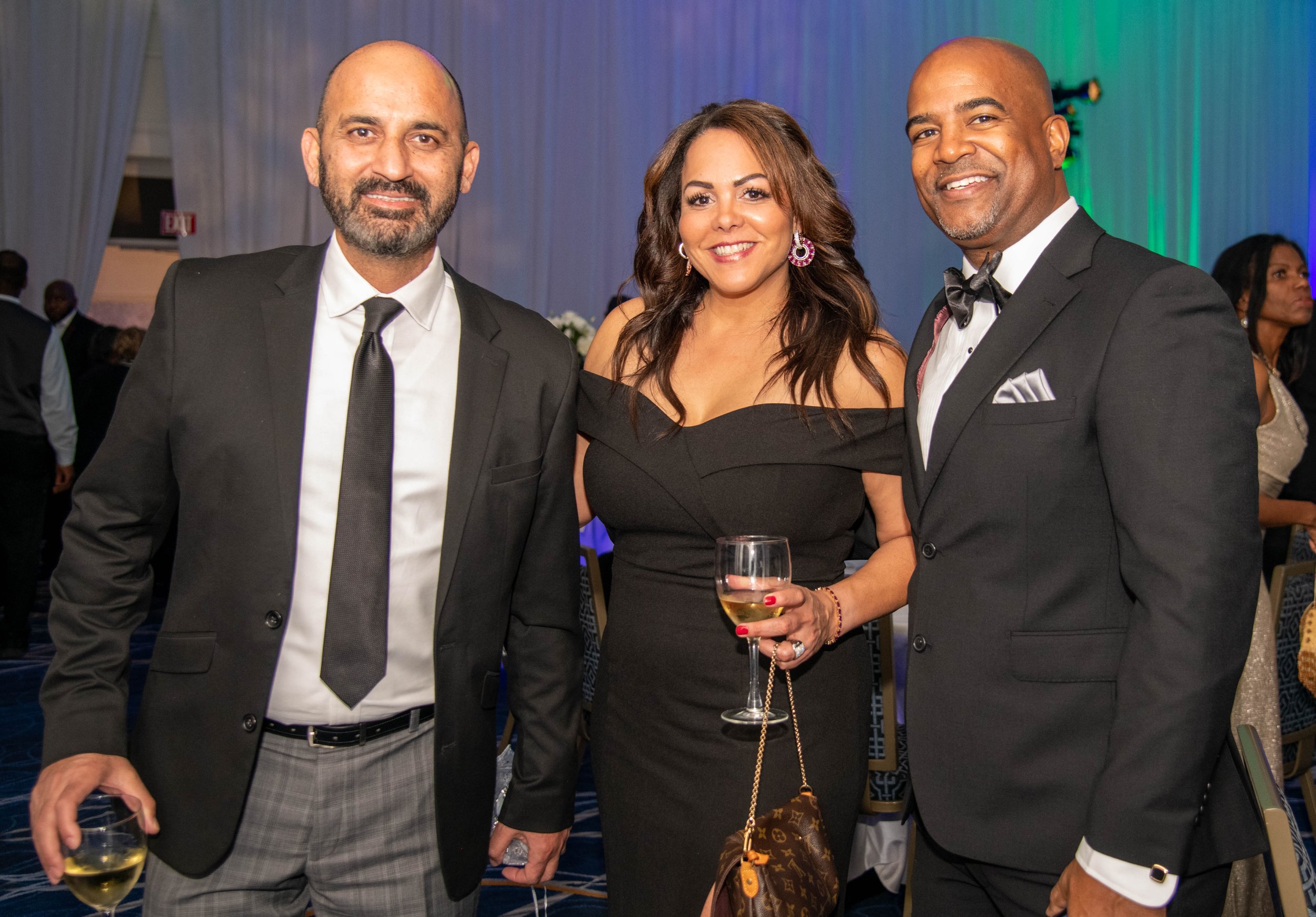 Three people dressed in formal attire at an indoor event, holding drinks, smiling at the camera.