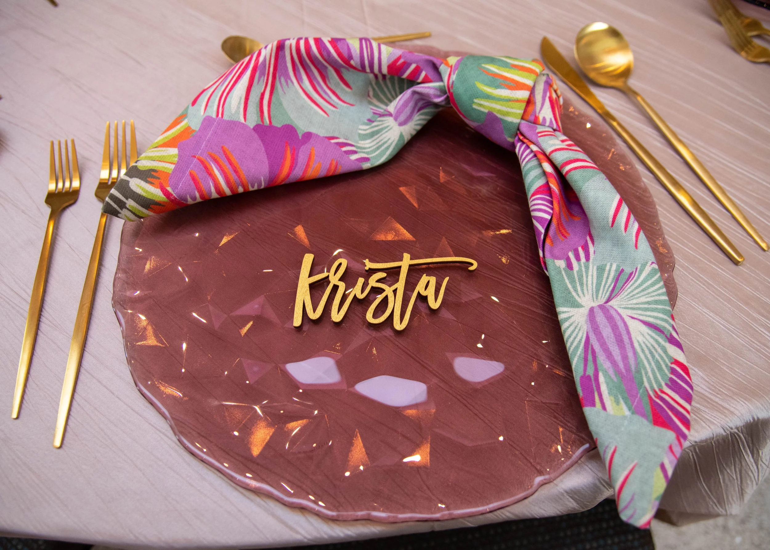 A table setting with a pink, faceted plate, gold utensils, a colorful floral napkin, and a name tag that says 'Krista'.