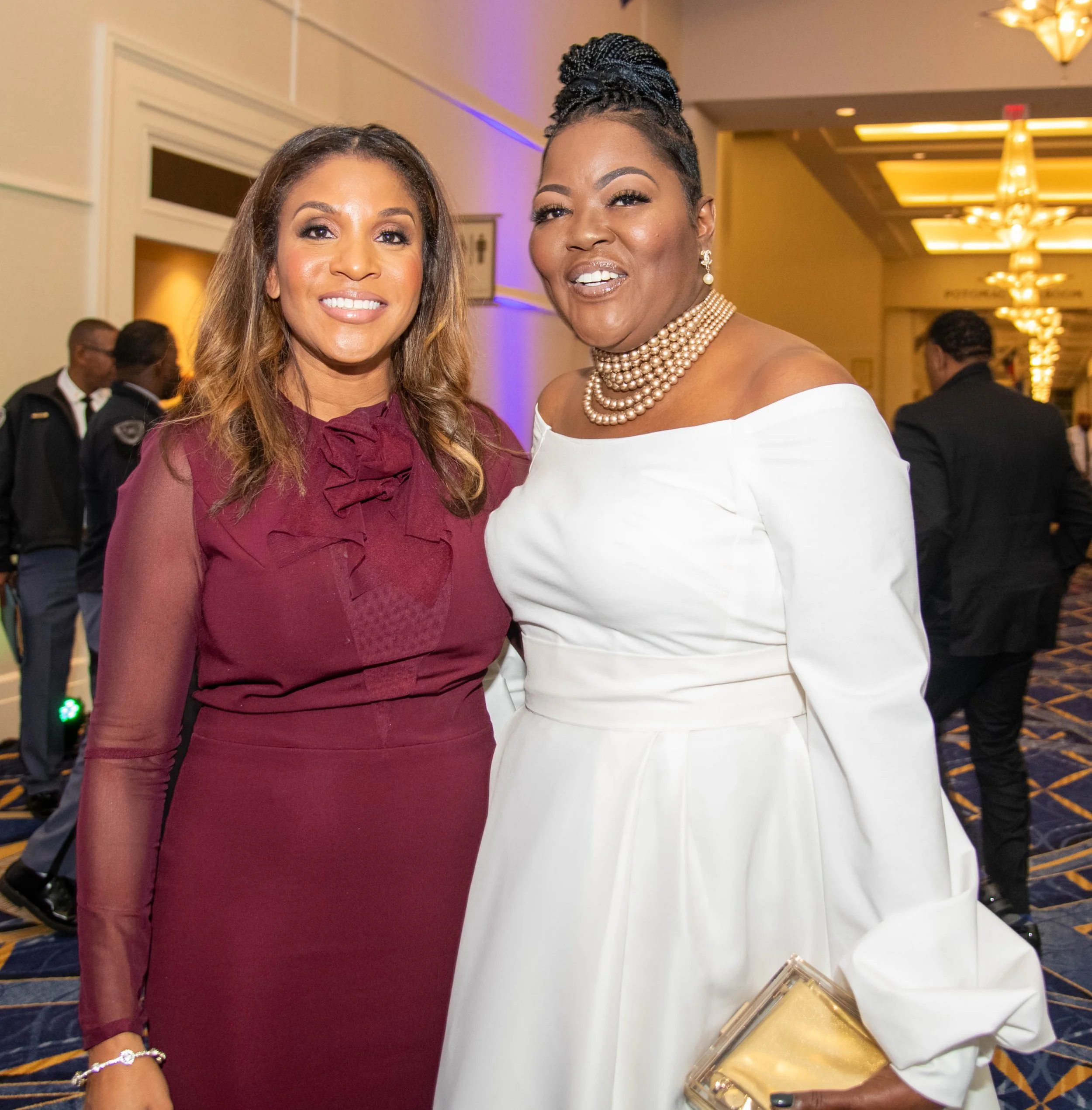 Two women smiling at an indoor event, one wearing a maroon dress with sheer sleeves and the other in a white off-shoulder dress with pearl jewelry, holding a gold clutch.