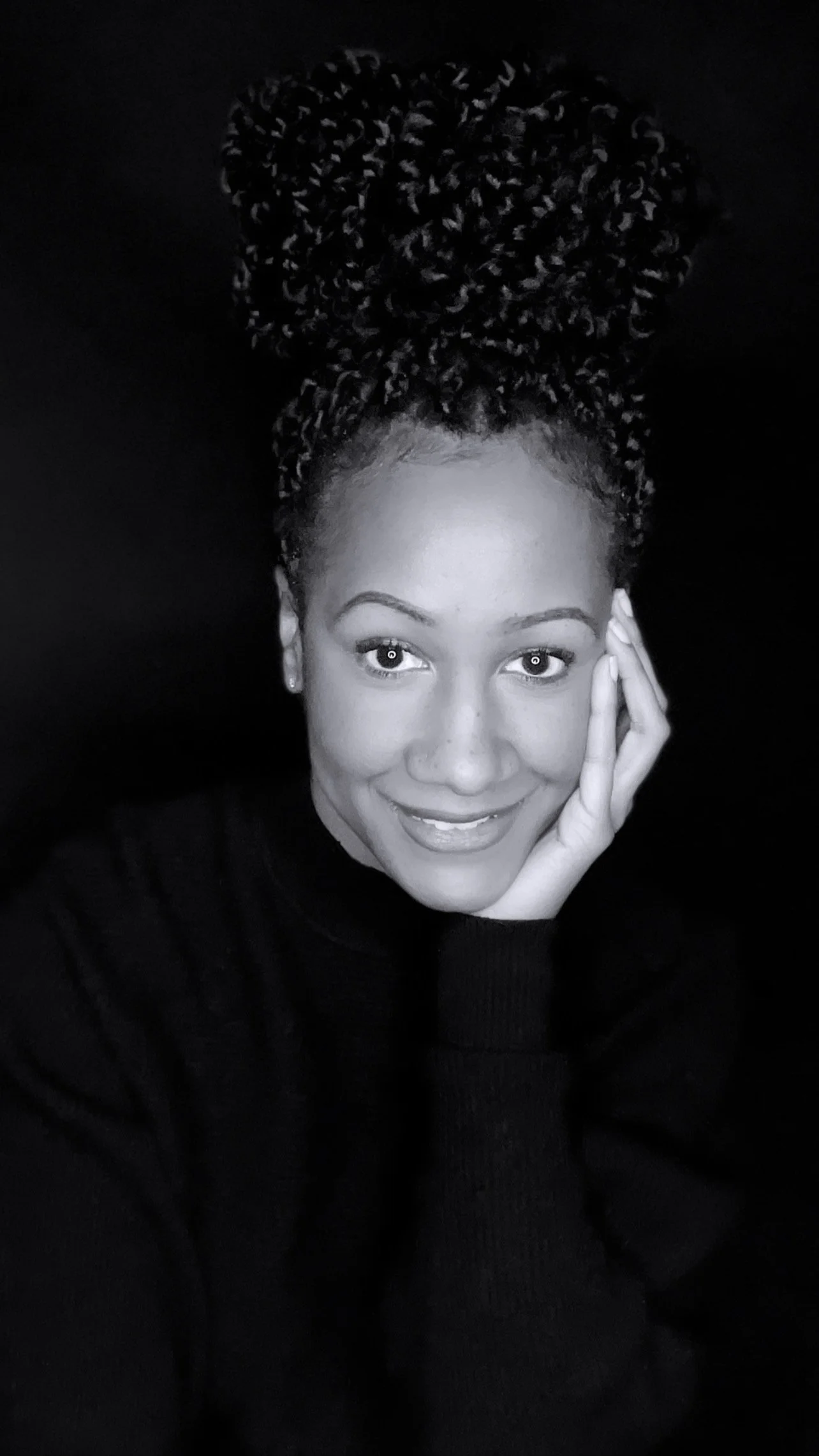 A smiling woman with curly hair styled in a high bun, wearing a dark top, resting her face on her hand against a black background.