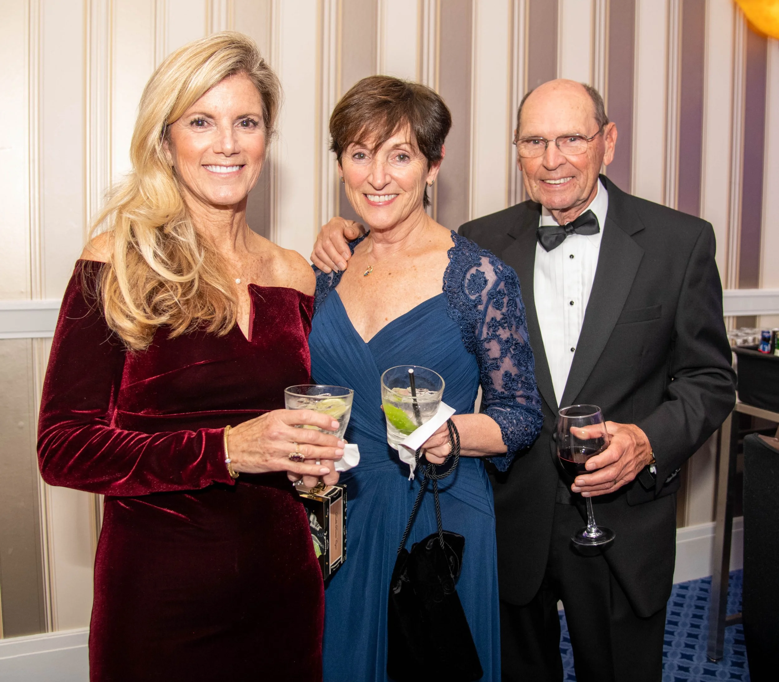 Three adults at a formal event, two women and one man, holding drinks and smiling at the camera.