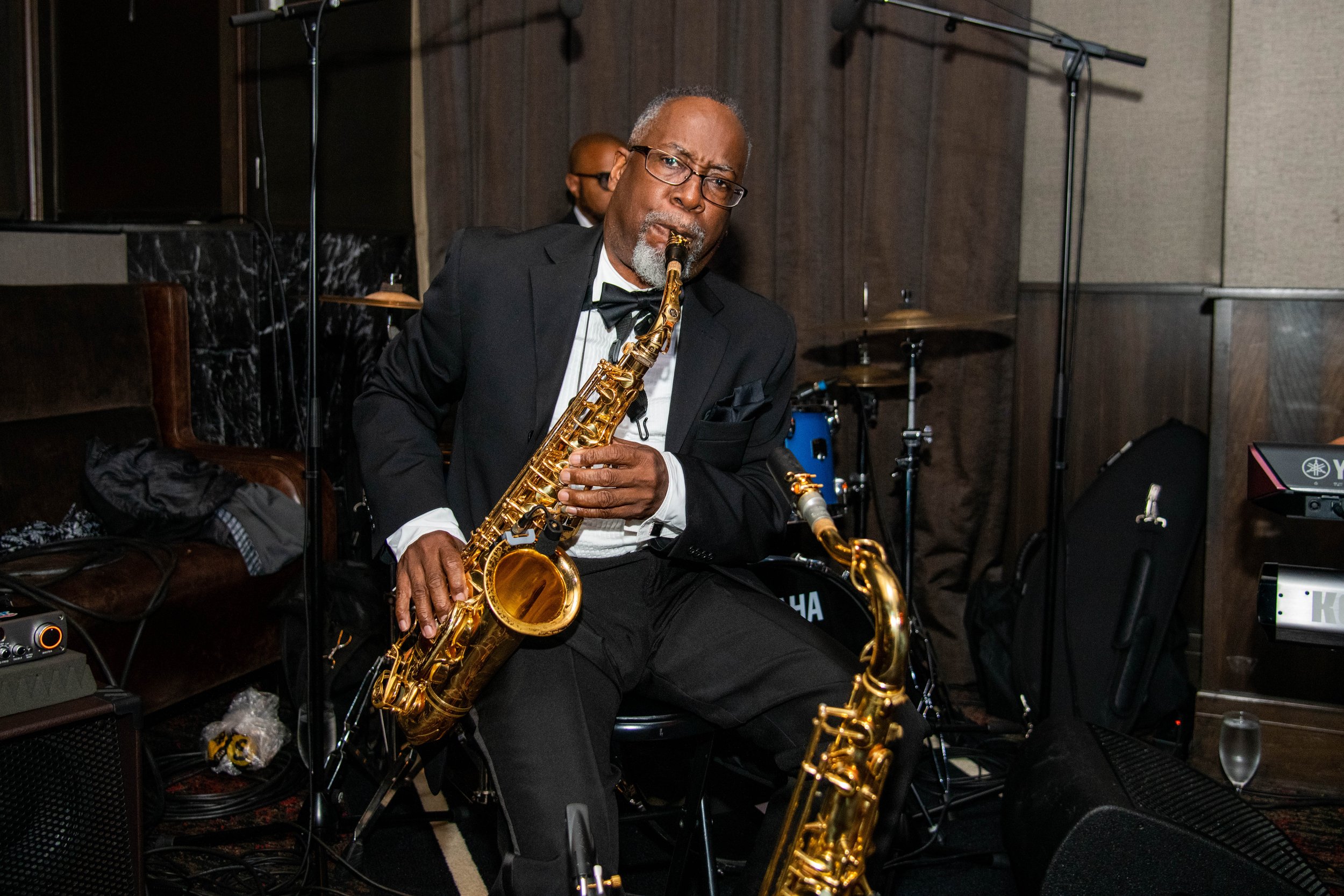 An older man in a tuxedo playing a gold saxophone during a performance indoors, with musical equipment and a small drum set in the background.