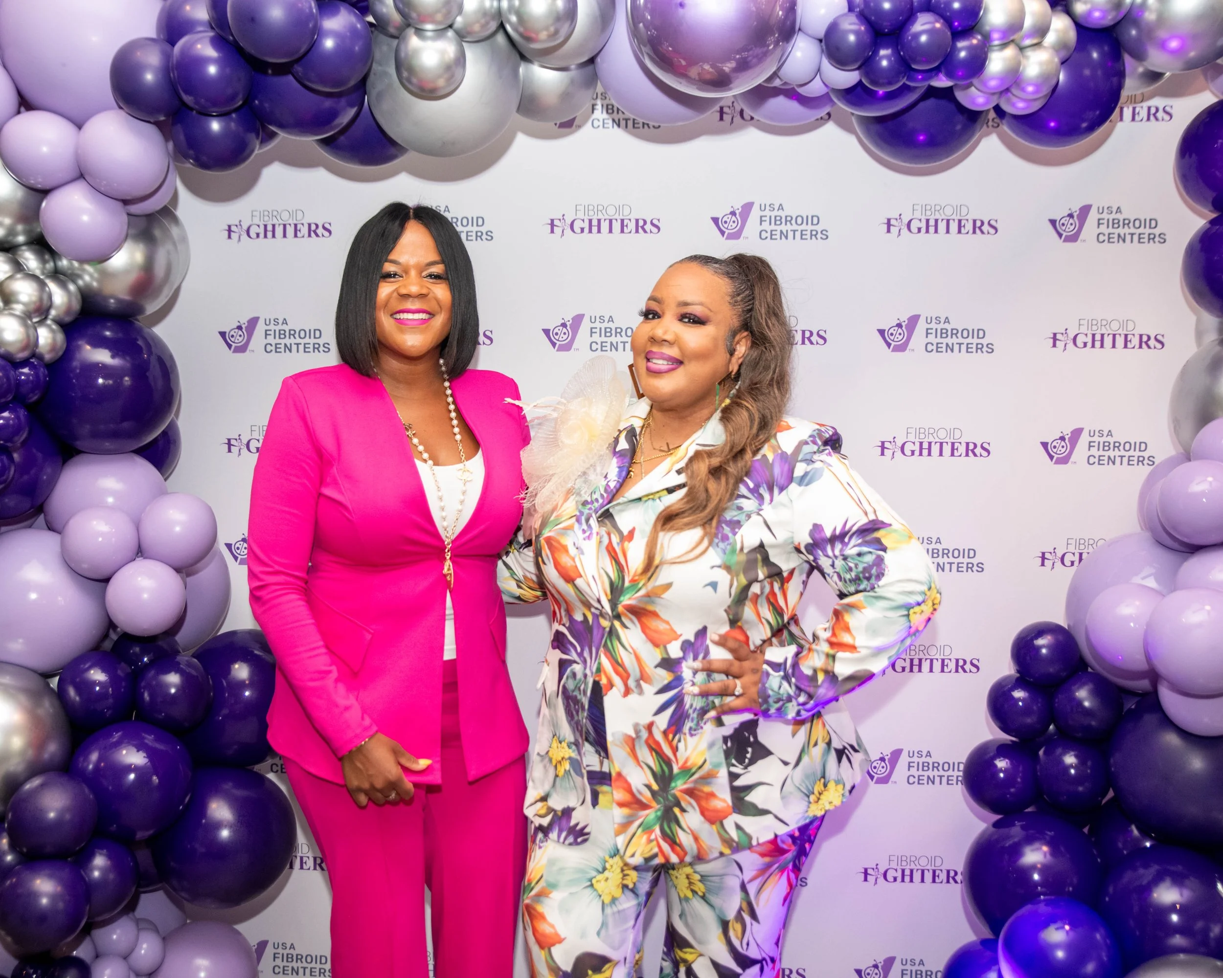 Two women standing in front of a backdrop with purple and silver balloons. The backdrop features the logos of USA Fibroid Centers and Fibroid Fighters. The woman on the left is wearing a bright pink suit with a white top and a pearl necklace, and the