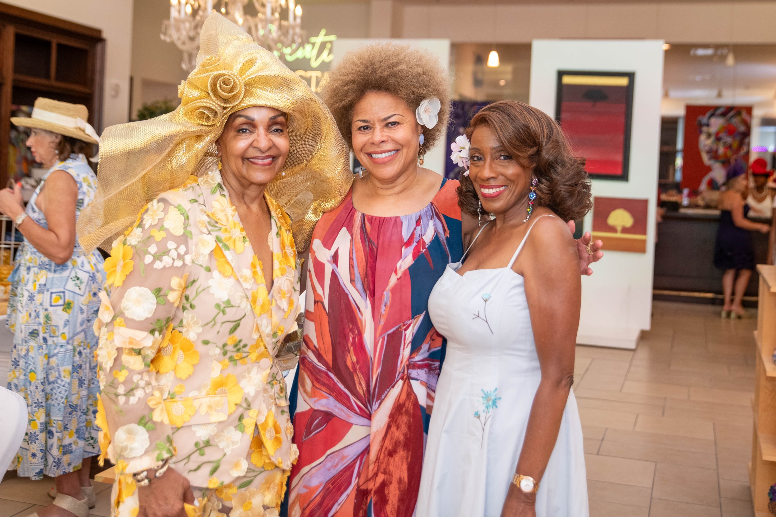 Three women posing together in an art gallery or boutique store, smiling. The woman on the left is wearing a floral dress with shades of yellow and beige, accessorized with a large, elaborate yellow hat. The woman in the middle is dressed in a colorf