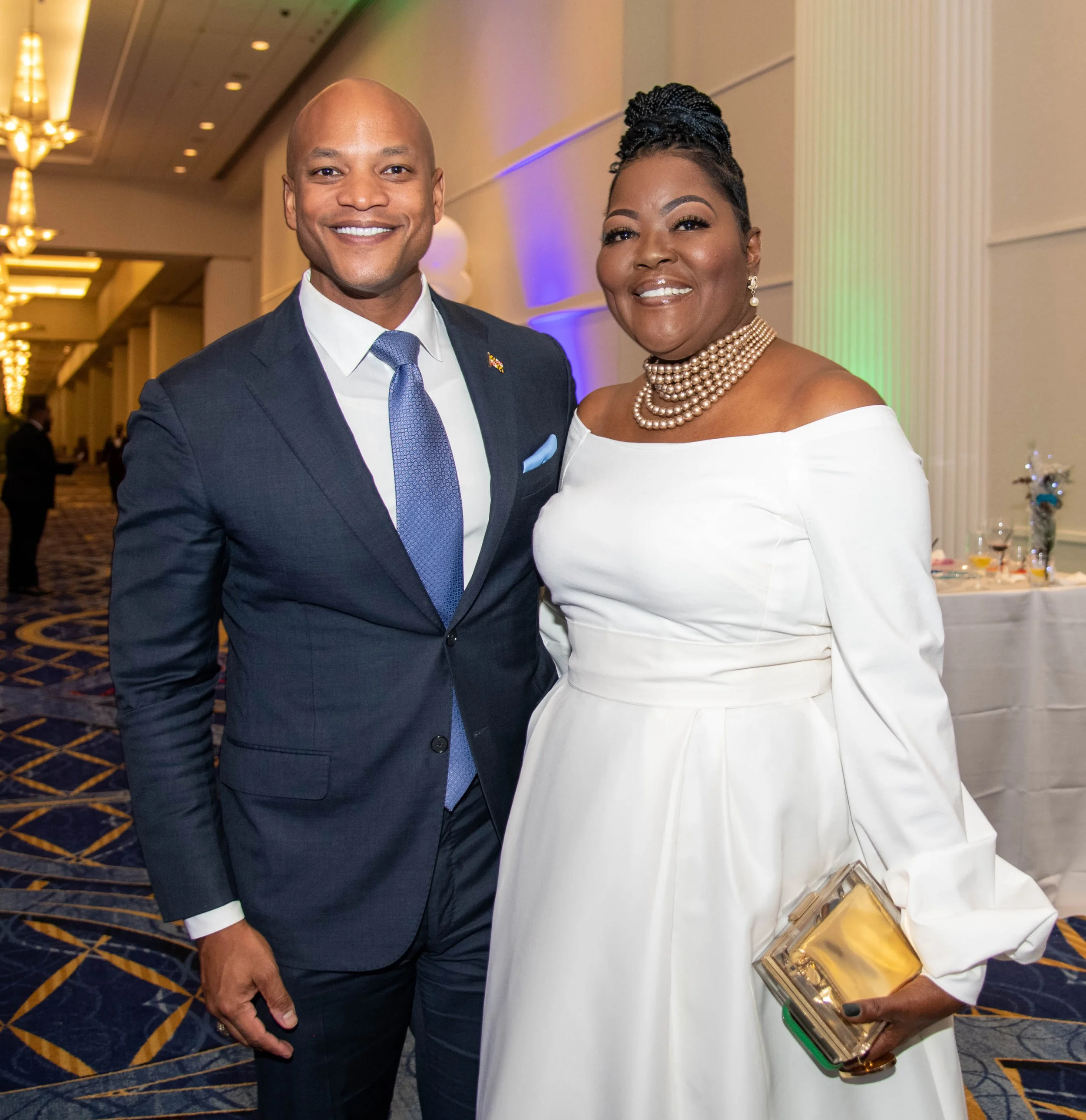A man and woman in formal attire attending a special event, smiling at the camera. The man is wearing a dark suit with a blue tie and ribbon pin, and the woman is wearing an elegant white dress with layered pearl necklaces, earrings, and holding a go