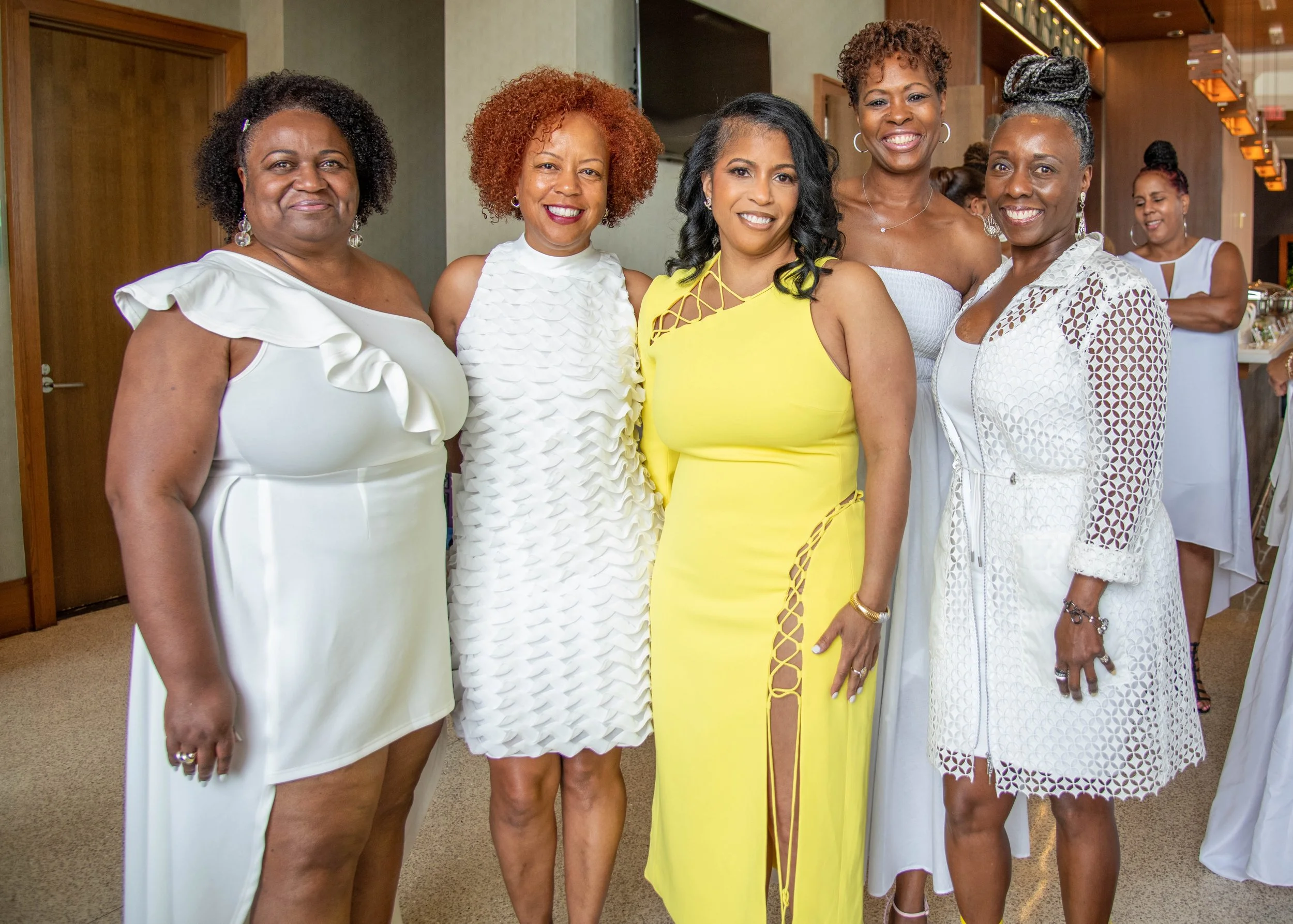 Six women in elegant dresses standing together inside a room, smiling at the camera at a social event or celebration.