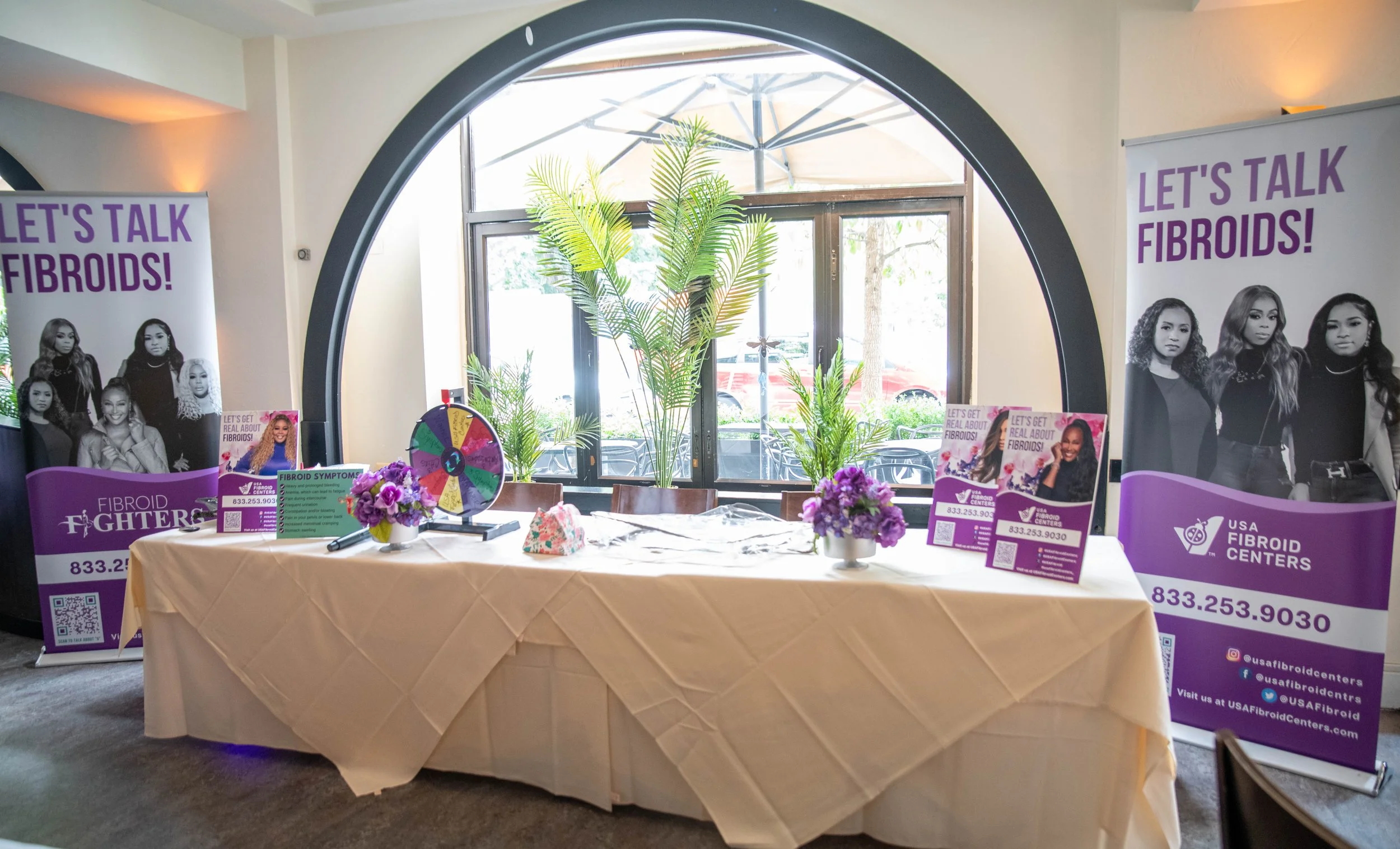 Table with purple flowers, informational flyers, and a prize wheel at a fibroids awareness event. Two large purple banners on either side display women and read 'Let's Talk Fibroids!' with contact information for USA Fibroid Centers. A window with pl