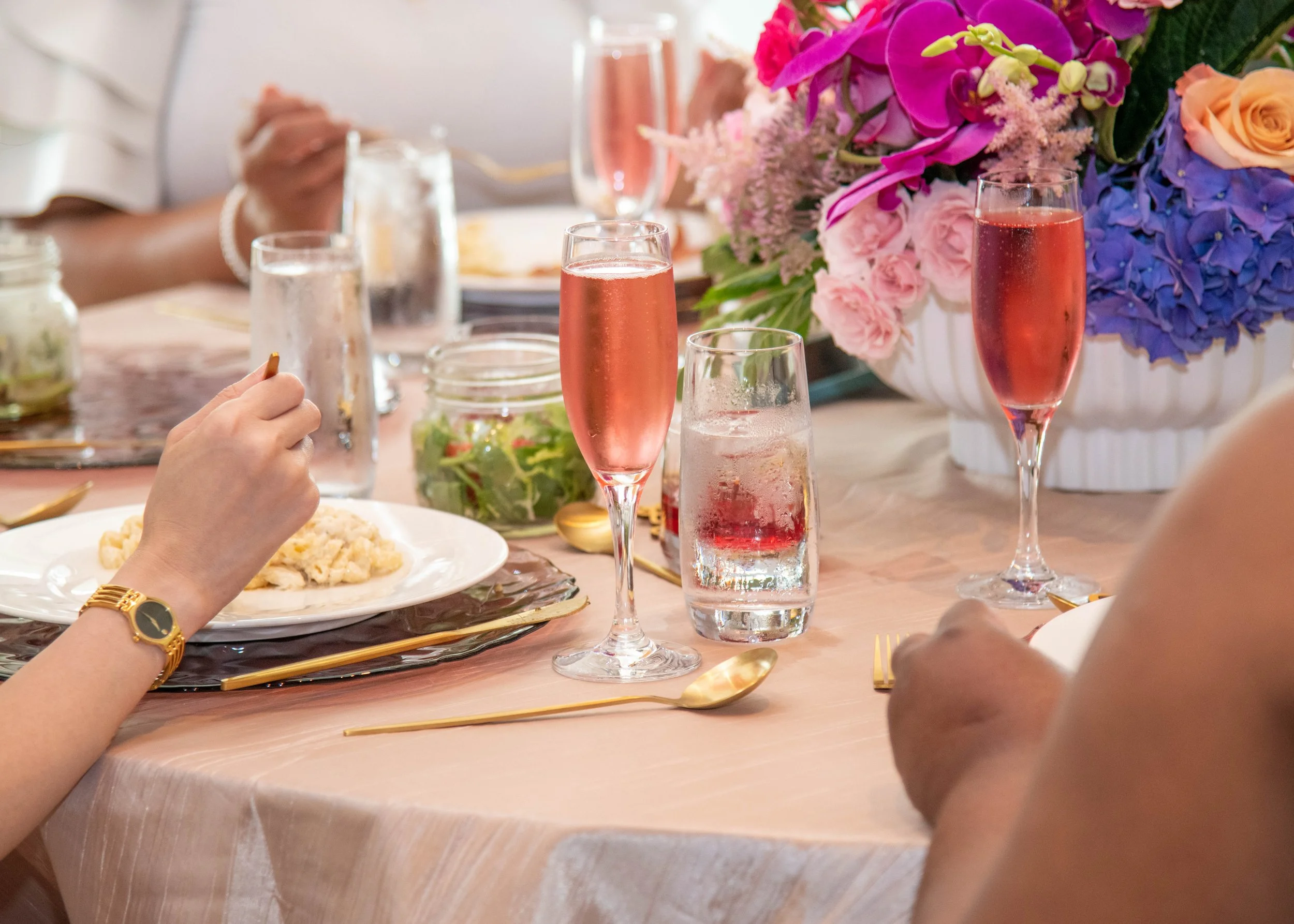 A table set for a celebration with pink and purple flowers, glasses of pink sparkling drinks, water, and a plate of food.