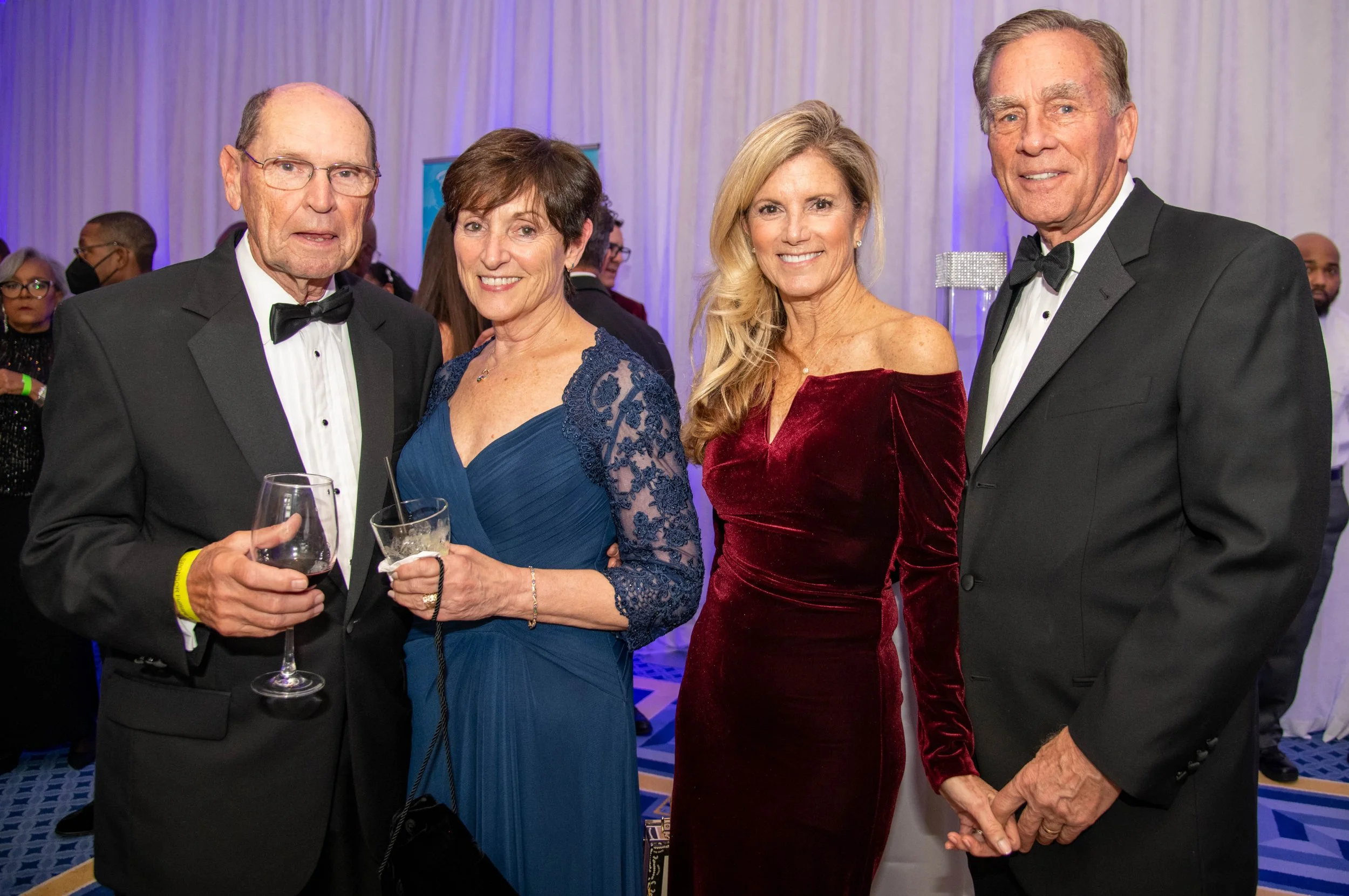 Four elegantly dressed adults at a formal event, standing together indoors, with a white curtain backdrop and purple lighting.