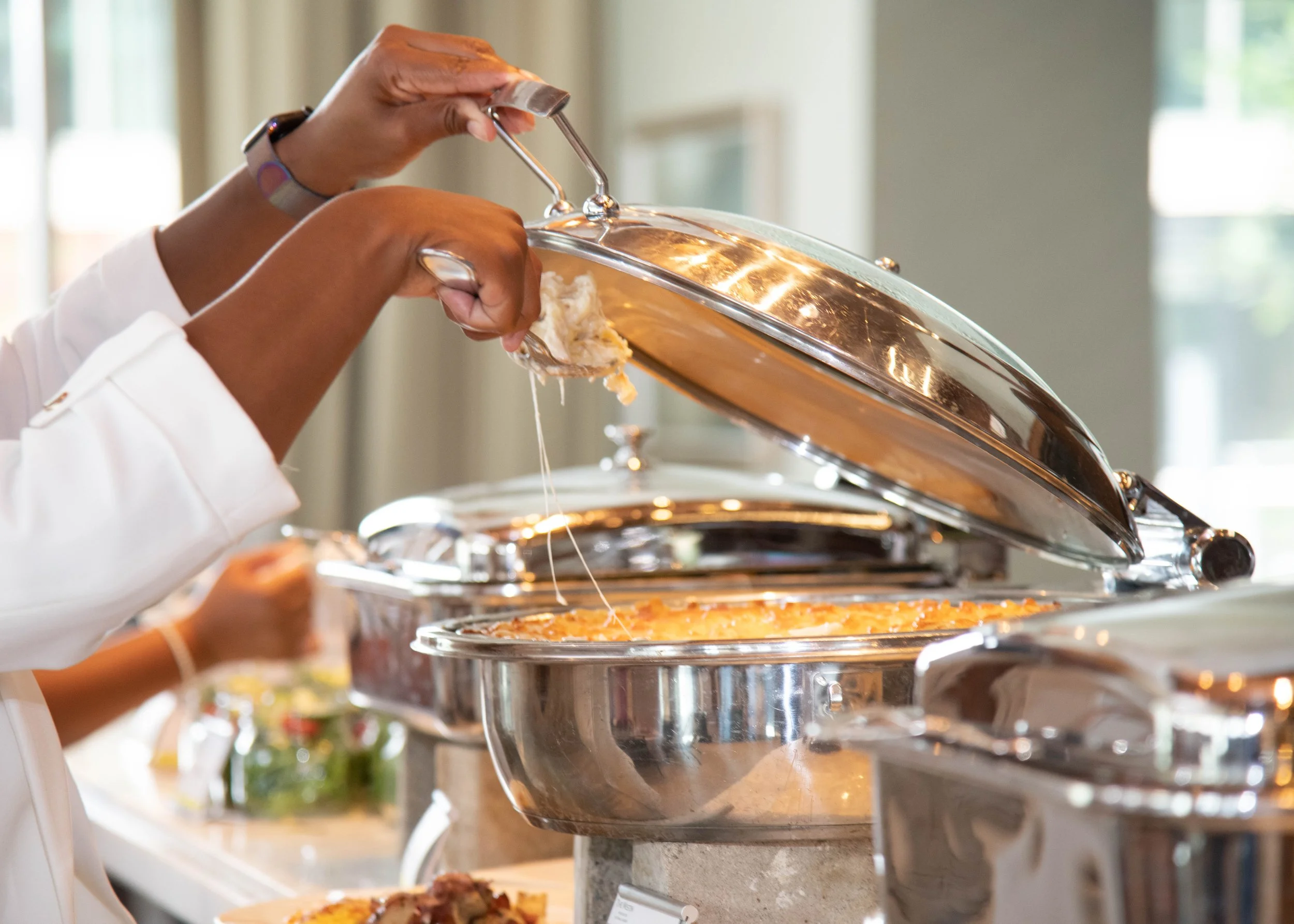 Two people are serving themselves food from chafing dishes at a buffet. One person is lifting the lid while the other waits nearby.