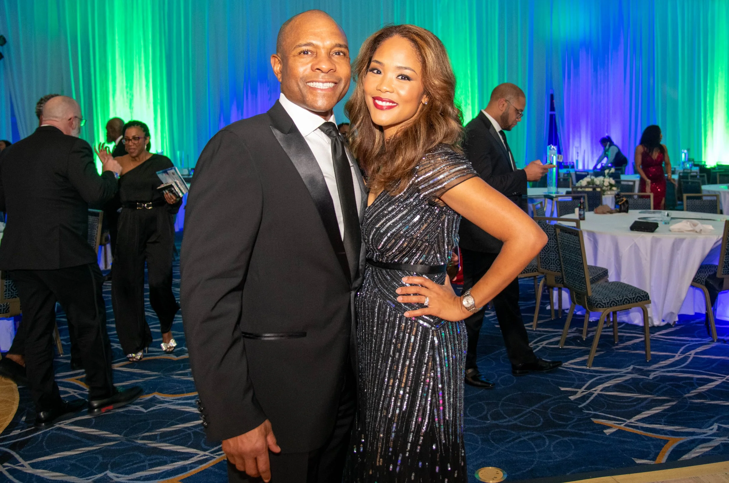 A smiling man in a black tuxedo and a woman in a sparkly black gown posing together at a formal event with other guests and green, blue, and purple lighting in the background.