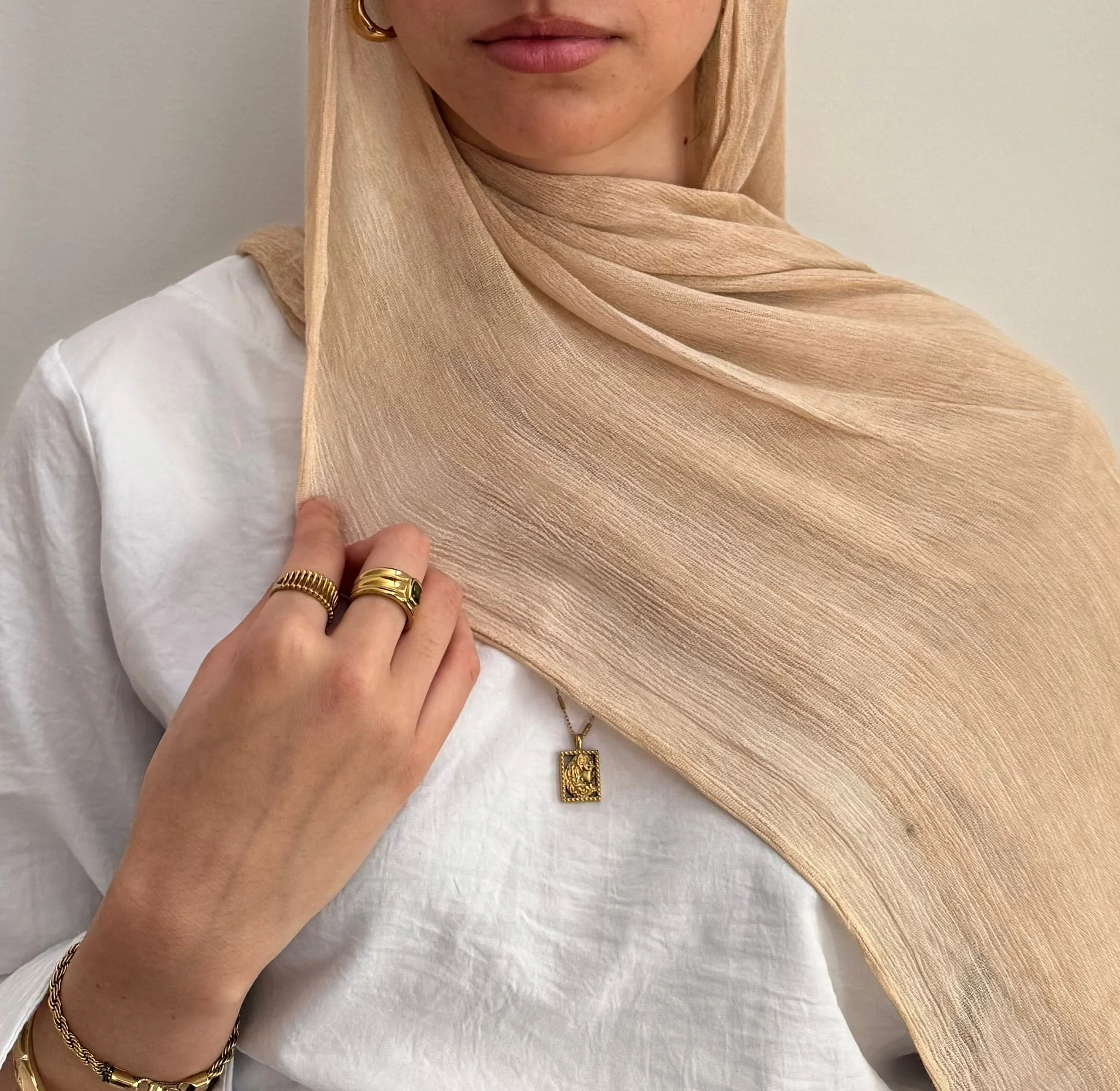 Woman wearing a beige hijab, white top, gold rings and jewelry, and a gold necklace, holding the hijab's edge with her hand.