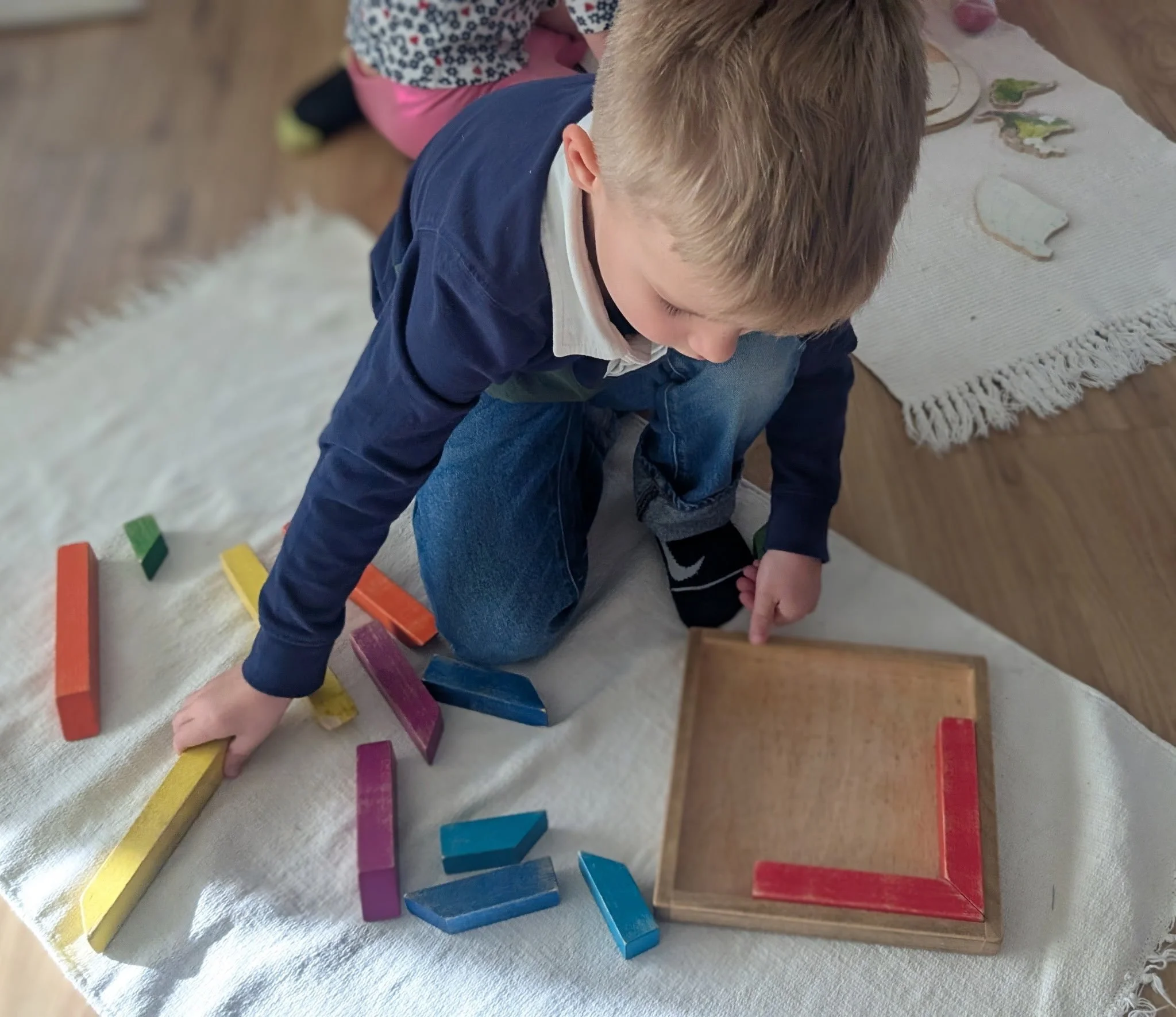 A young boy taps red tiles on a wooden frame resembling a square, part of a colorful tower game, with scattered colored tiles on a white cloth and a woman in the background.