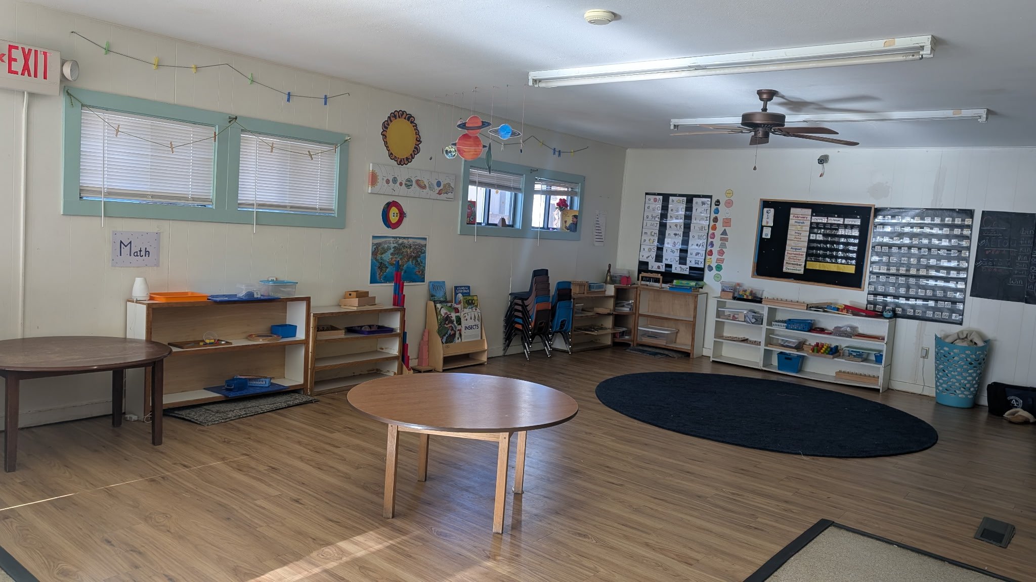 Empty preschool classroom with wooden floors, small tables, shelving units, educational posters, and classroom supplies along the walls.