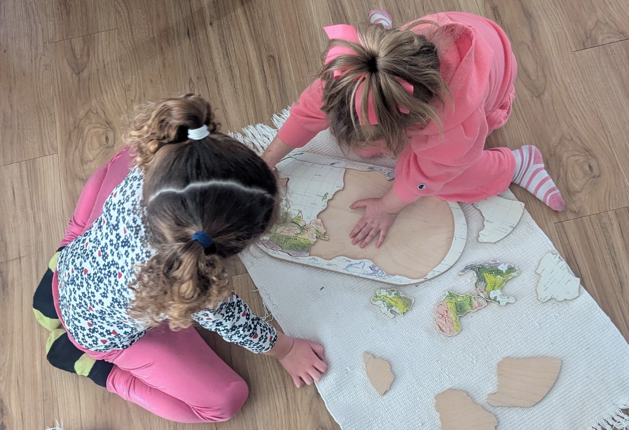 Two young girls are working together on a wooden puzzle of a map placed on a white cloth on a wooden floor.