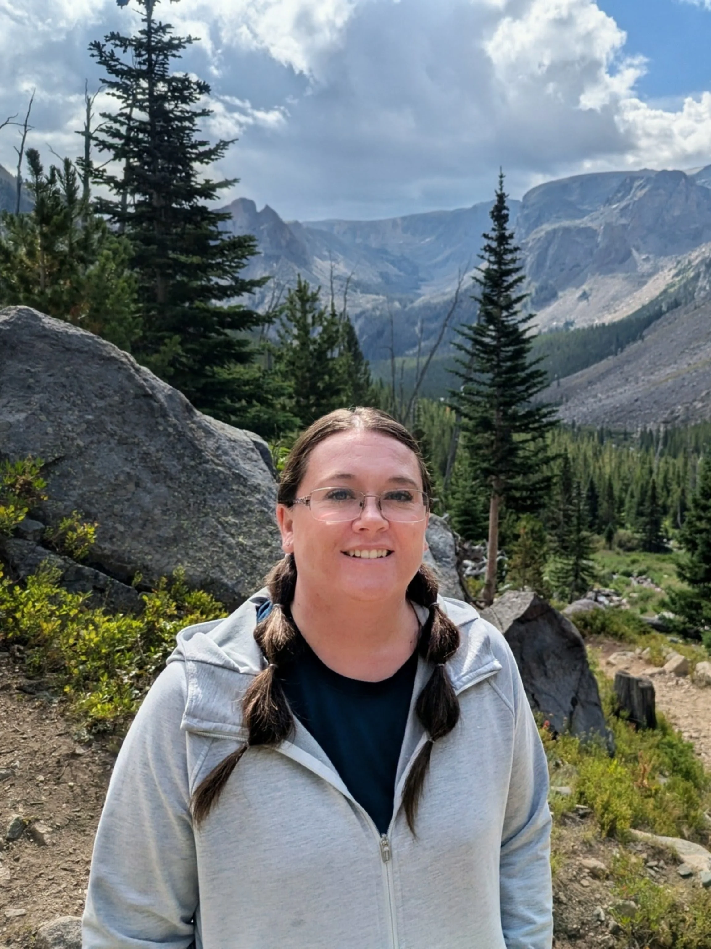 A woman with glasses and pigtails smiling in a mountainous forest landscape with pine trees, large rocks, and mountains in the background under a partly cloudy sky.