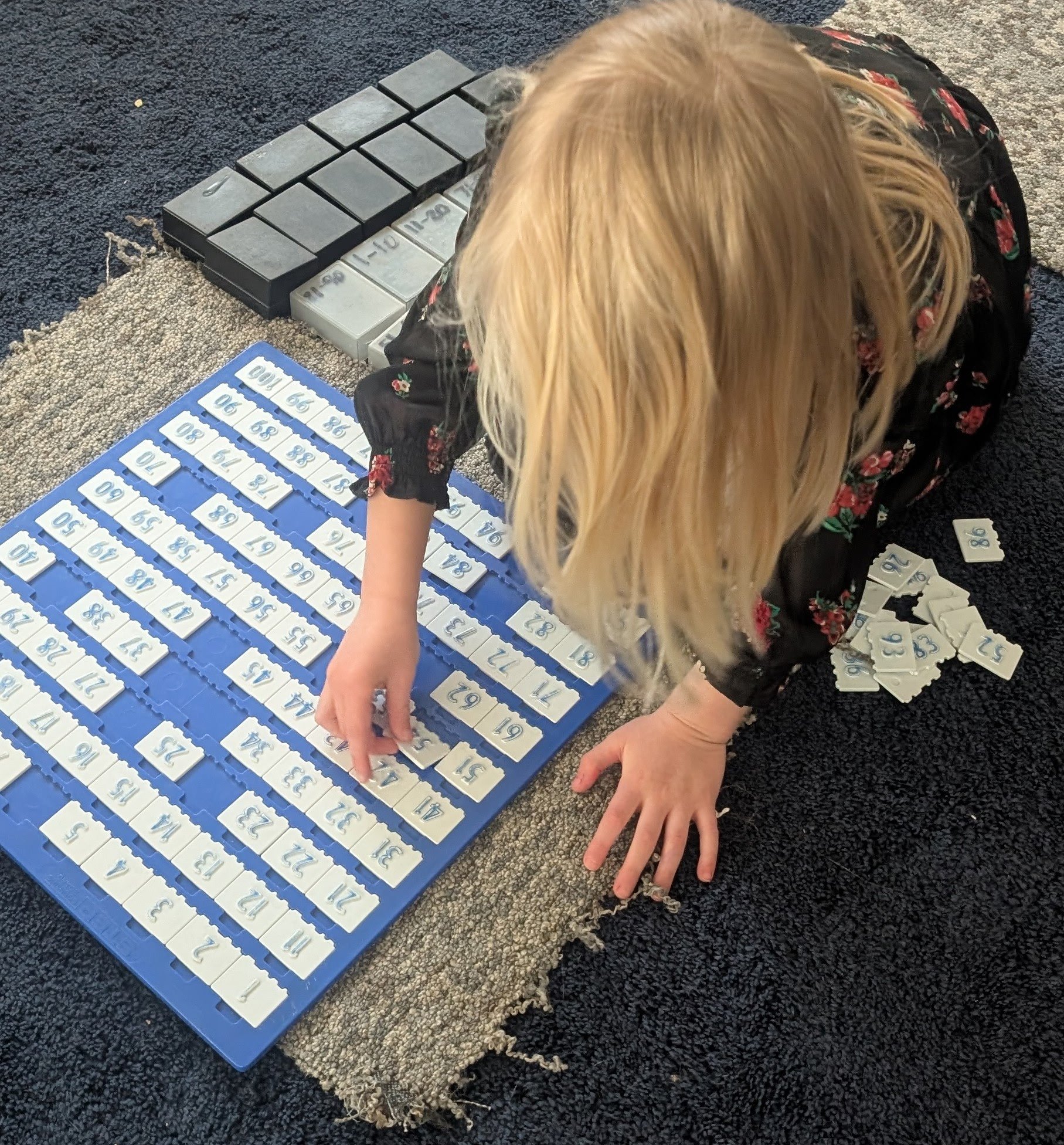 A young girl with long blonde hair is sitting on a carpeted floor, organizing dominoes on a blue game board. There are stacks of dominoes nearby, and the girl is reaching for a domino with the number 50.