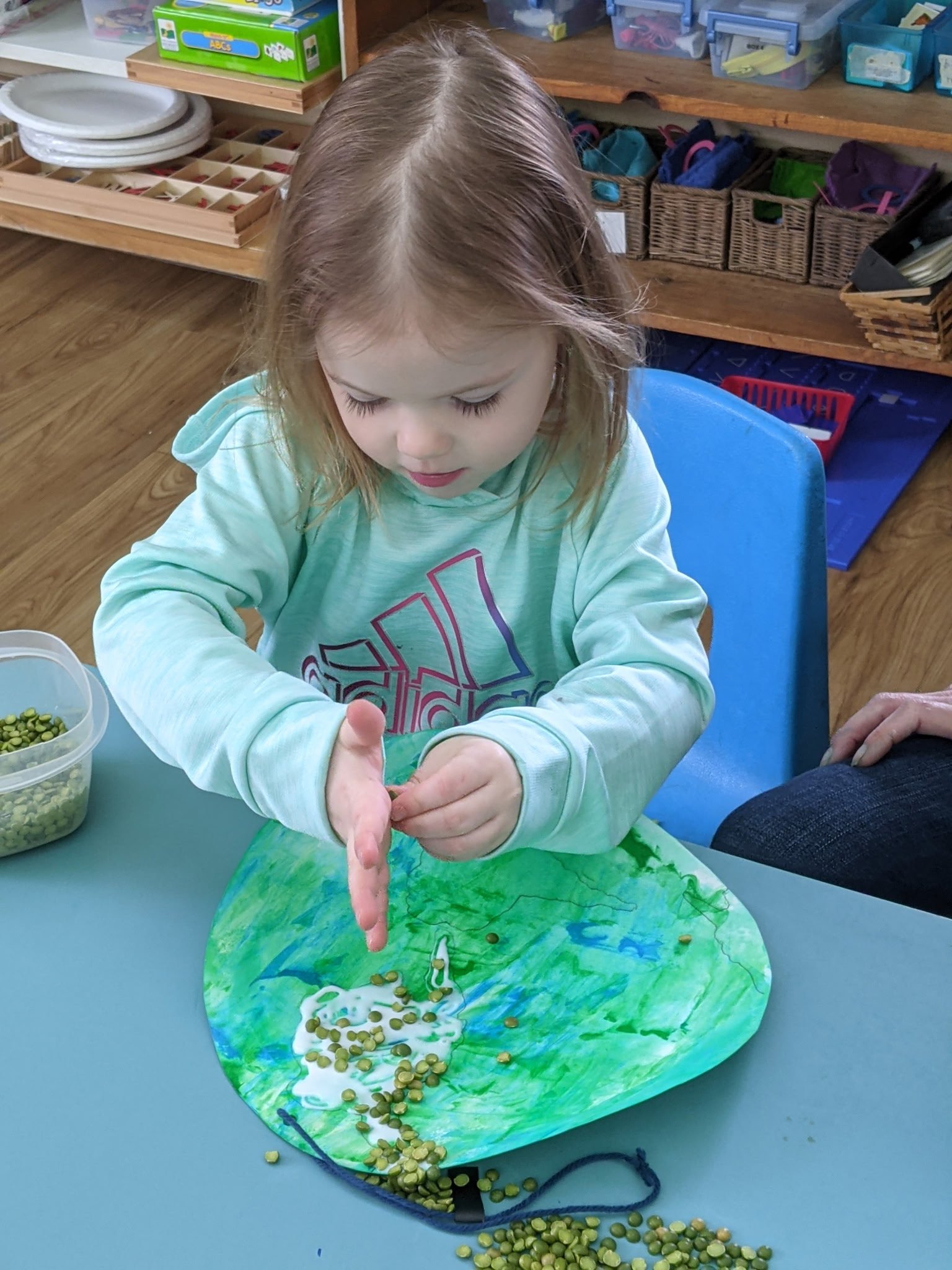 A young girl with light brown hair sits at a table, focusing on her craft project with green lentils and glue on a painted paper plate.