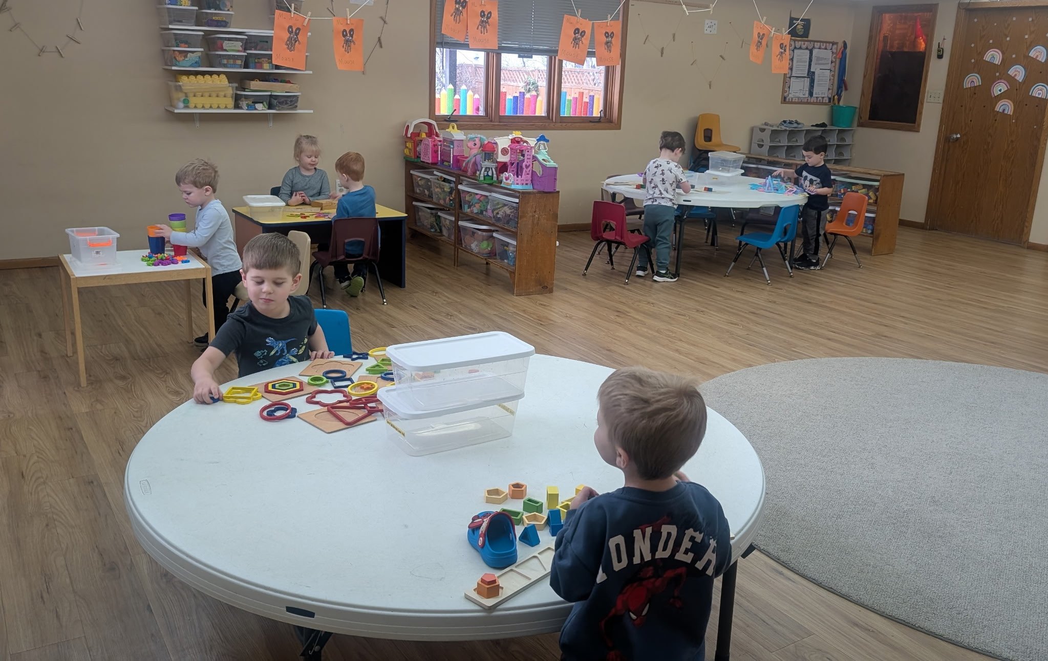 Six children playing with toys and engaging in activities in a preschool classroom with wooden floors, shelves of toys, and colorful decorations.
