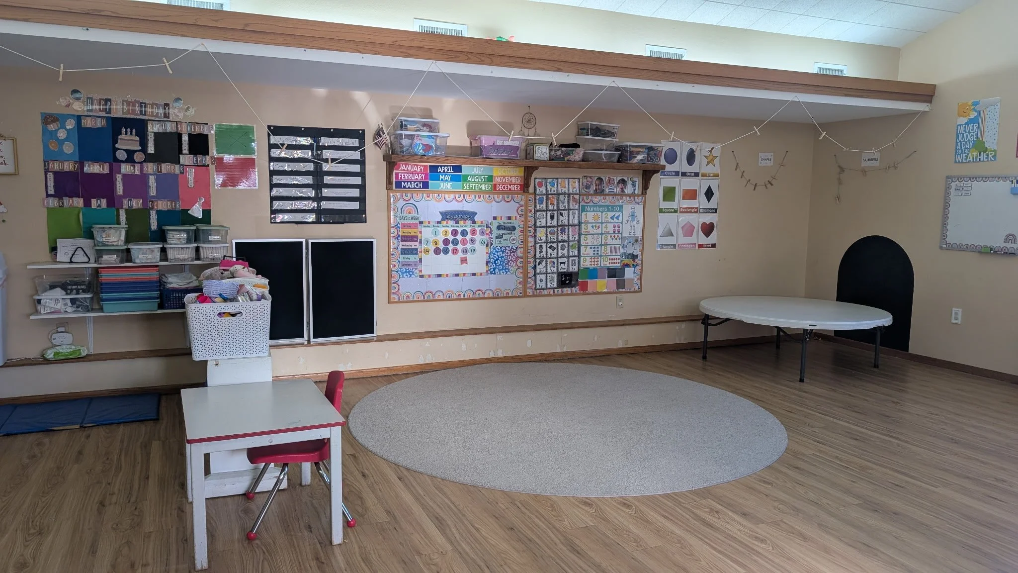 Empty classroom with children's educational posters, a small white table with a red chair, and a round gray rug on a wood floor.