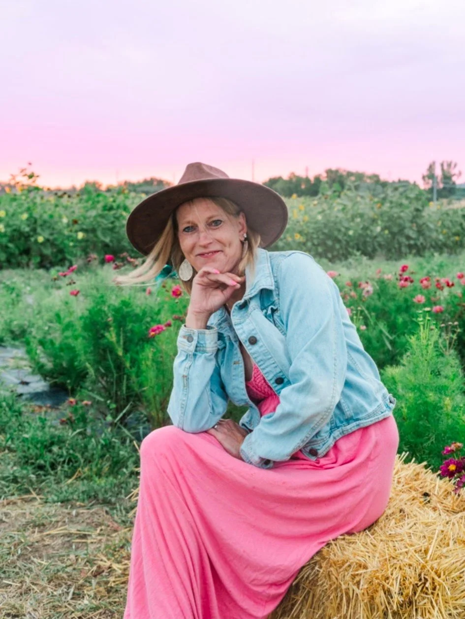 A woman with blonde hair, wearing a wide-brimmed hat, denim jacket, pink dress, and earrings, sitting on a hay bale in a flower field during sunset.