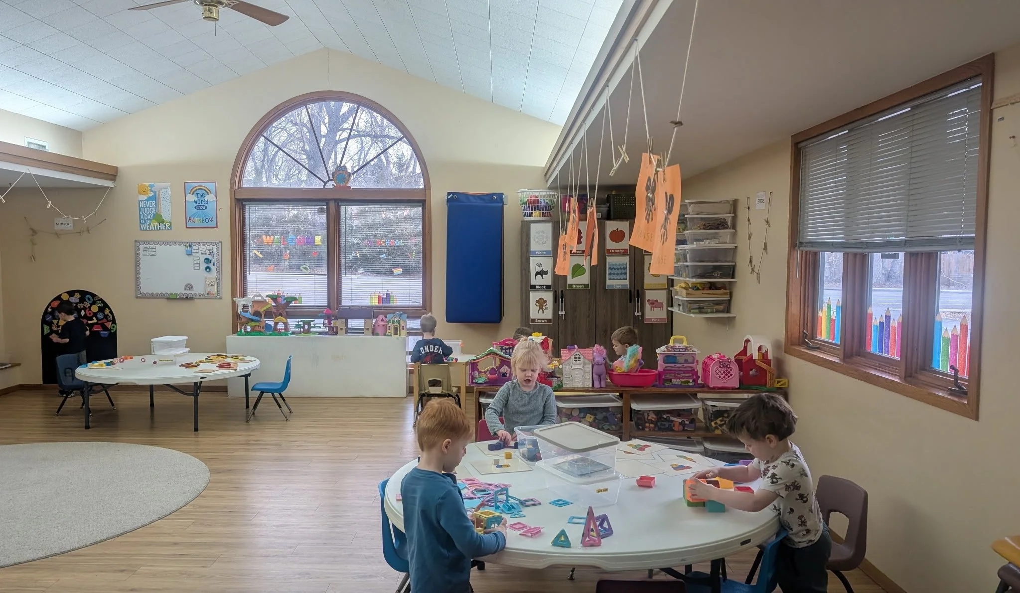 Children playing and working with toys in a colorful classroom with large arched windows and educational posters.